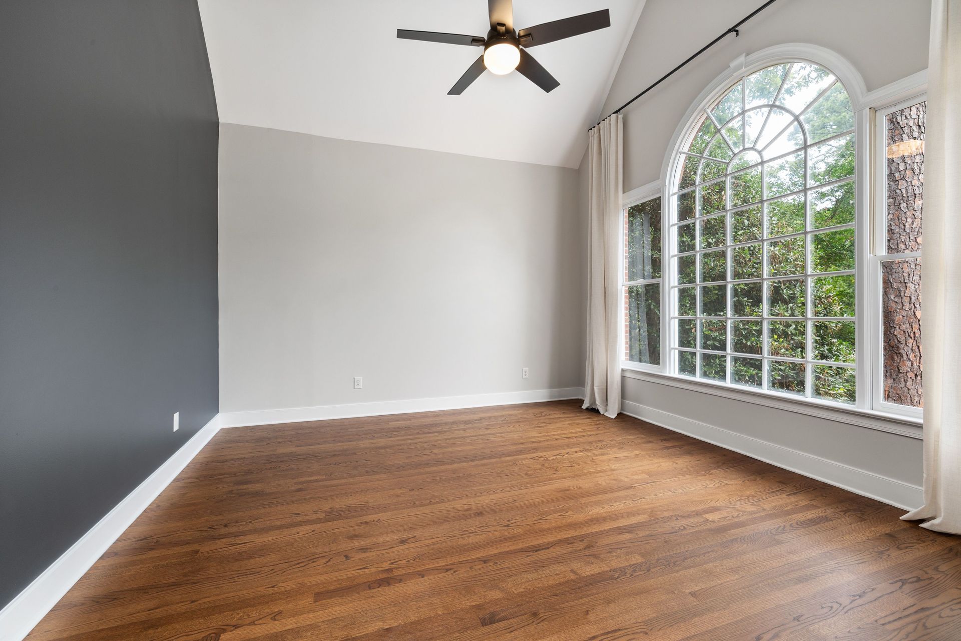 Empty room with hardwood floor, gray walls, vaulted ceiling, and large arched windows.