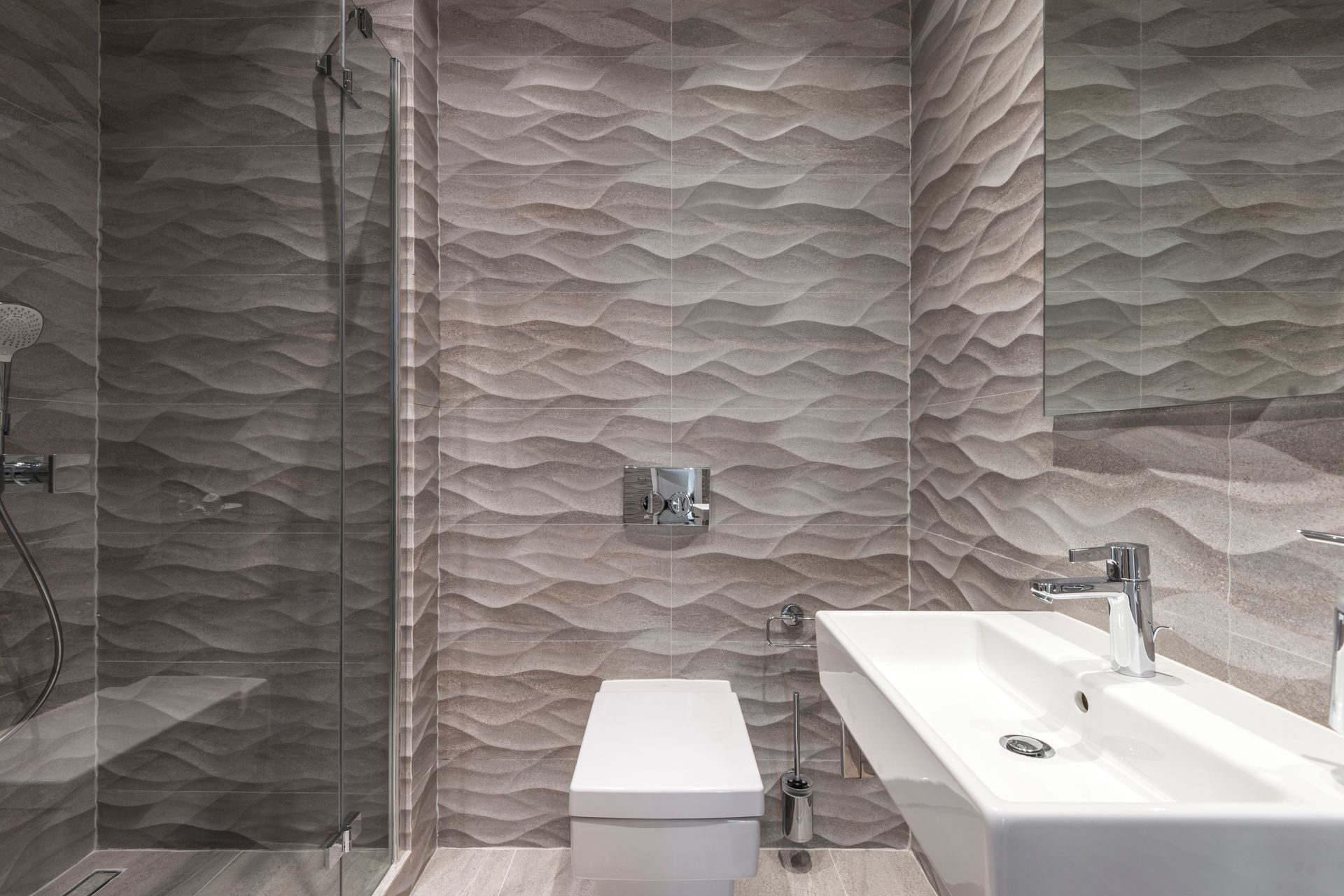 Modern bathroom with textured gray tile, a toilet, and a white sink beside a glass shower.
