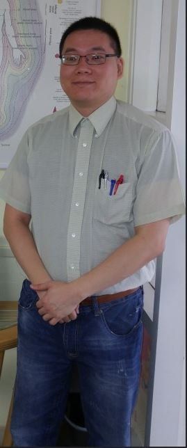 Person standing indoors in a light green shirt and jeans, hands clasped, with pens in the pocket.