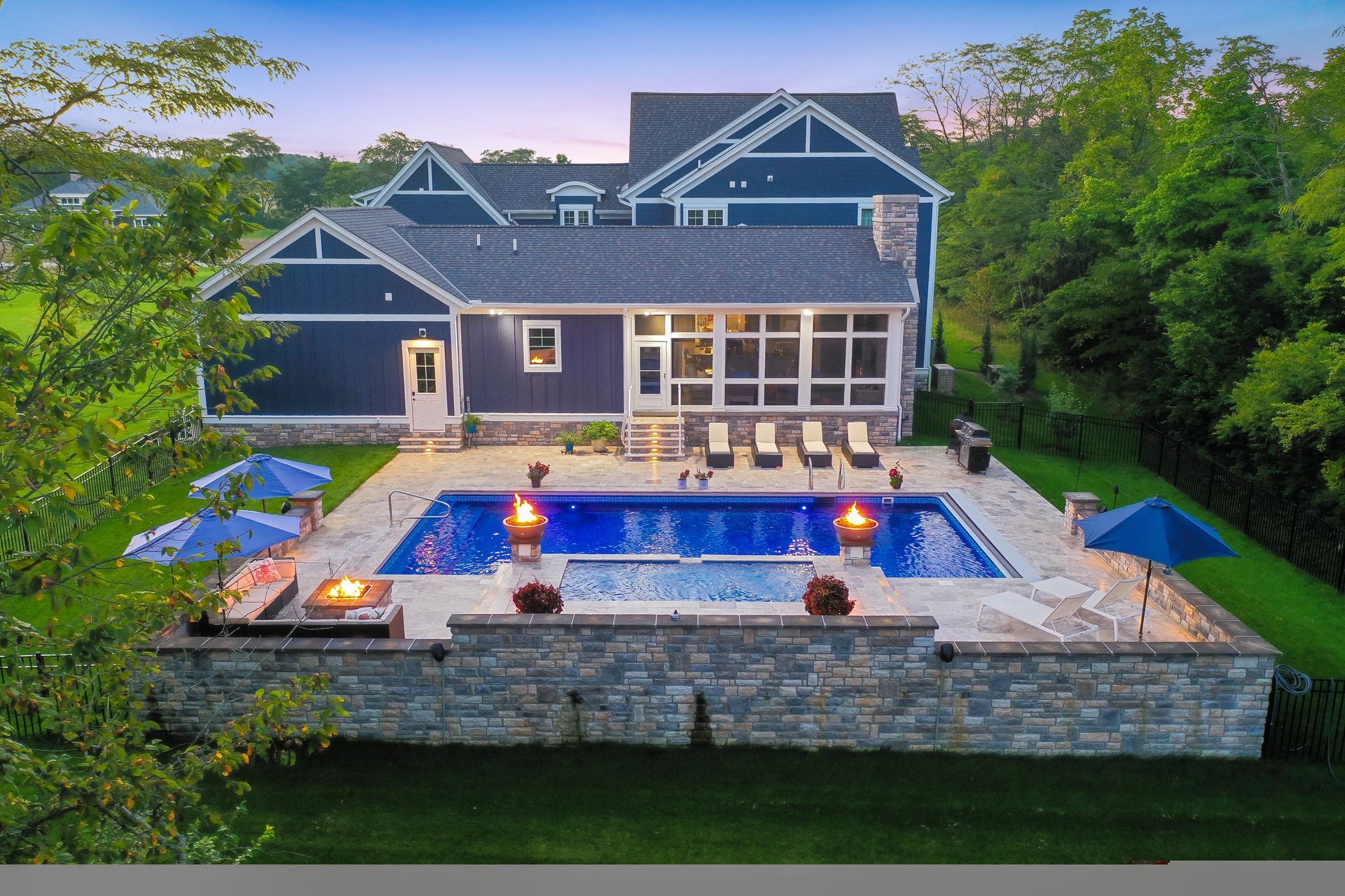 Modern gray house with lit pool and patio at dusk, surrounded by lush greenery