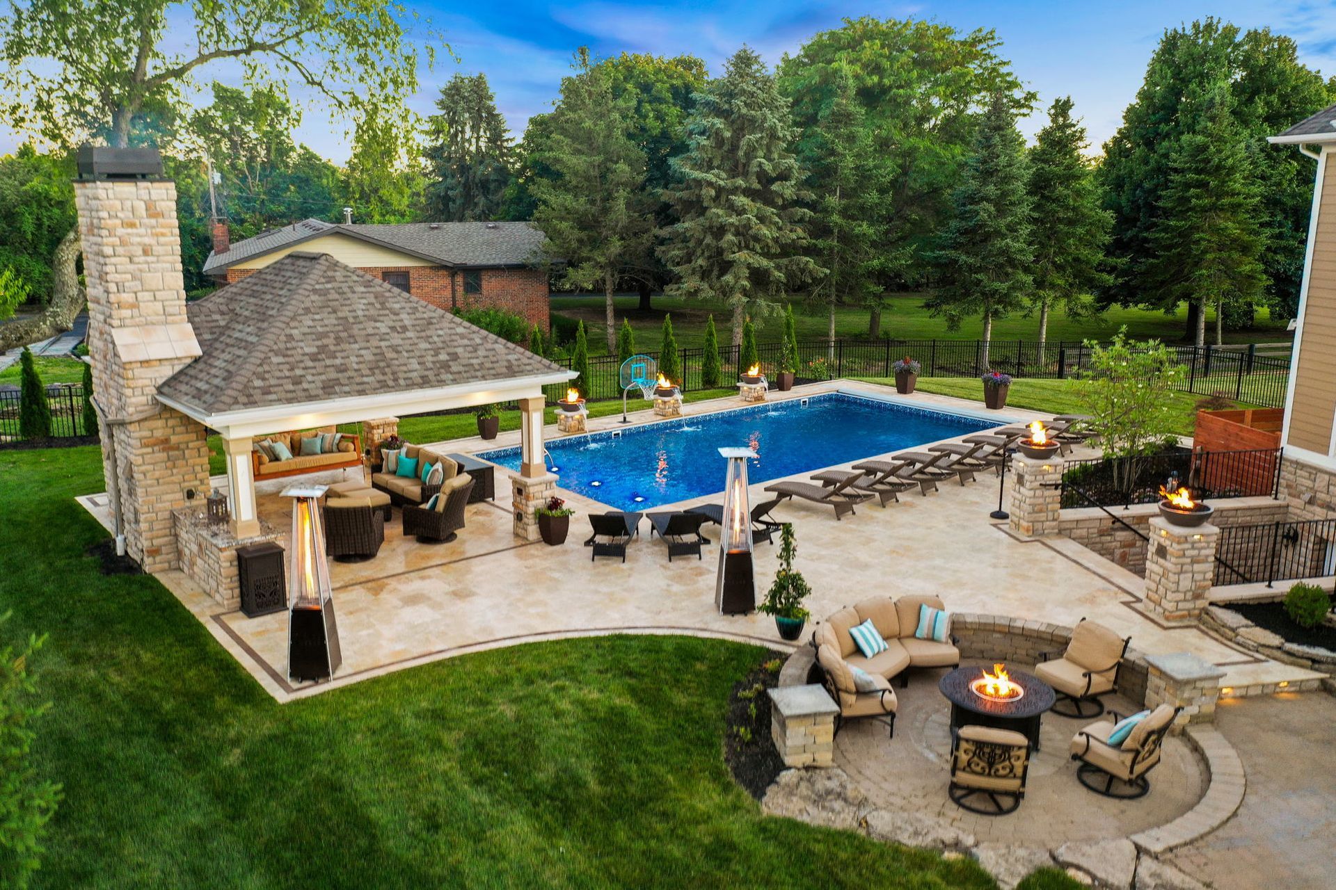 Backyard pool patio with stone fireplace, lounge chairs, and green lawn surrounded by trees