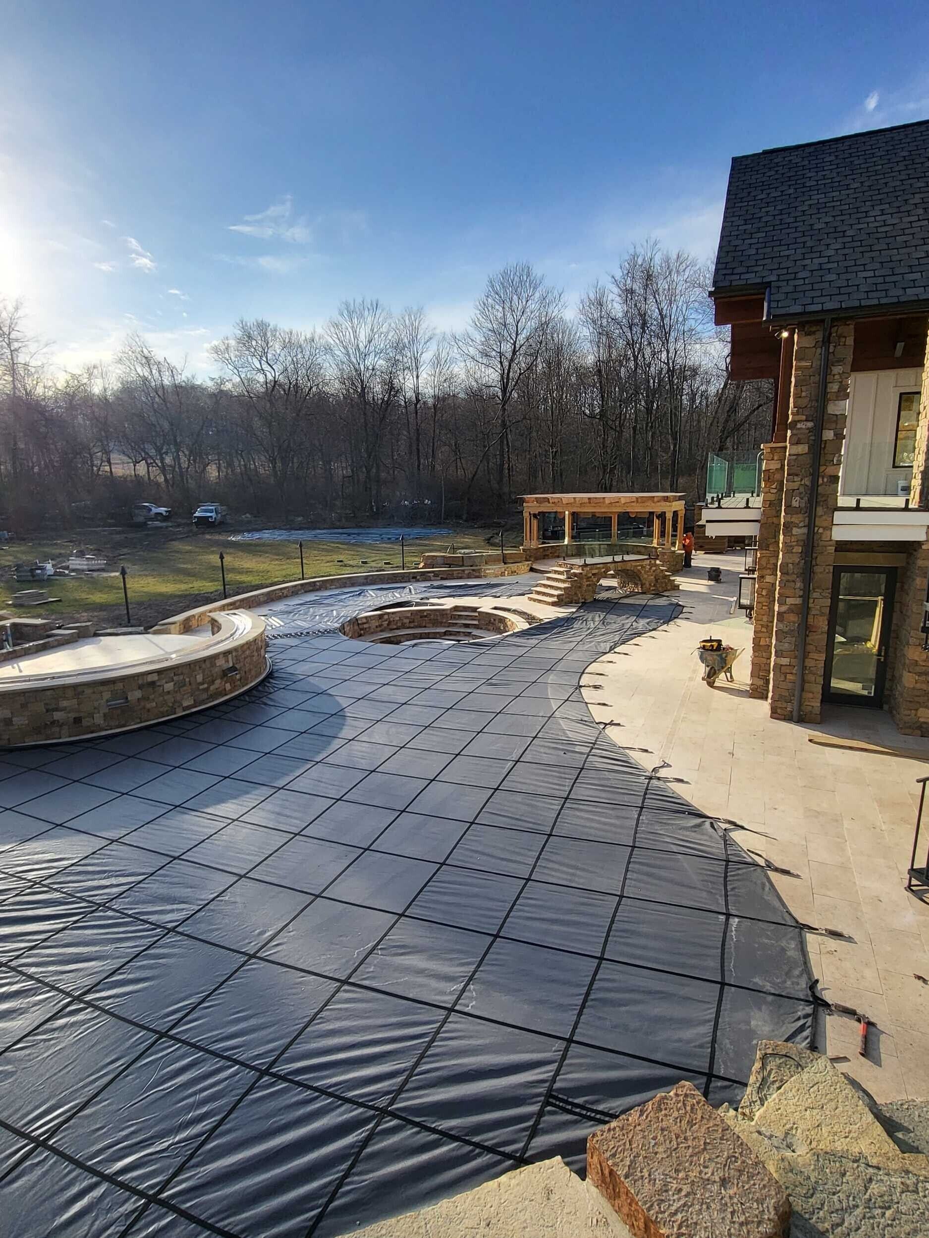 Backyard patio under construction with black tarped ground, stone fire pit, and wooden deck by a house