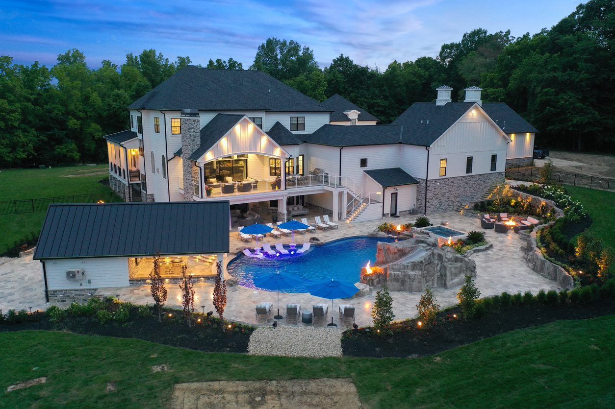 Luxury home with lit pool and stone patio at dusk, surrounded by trees and landscaped grounds