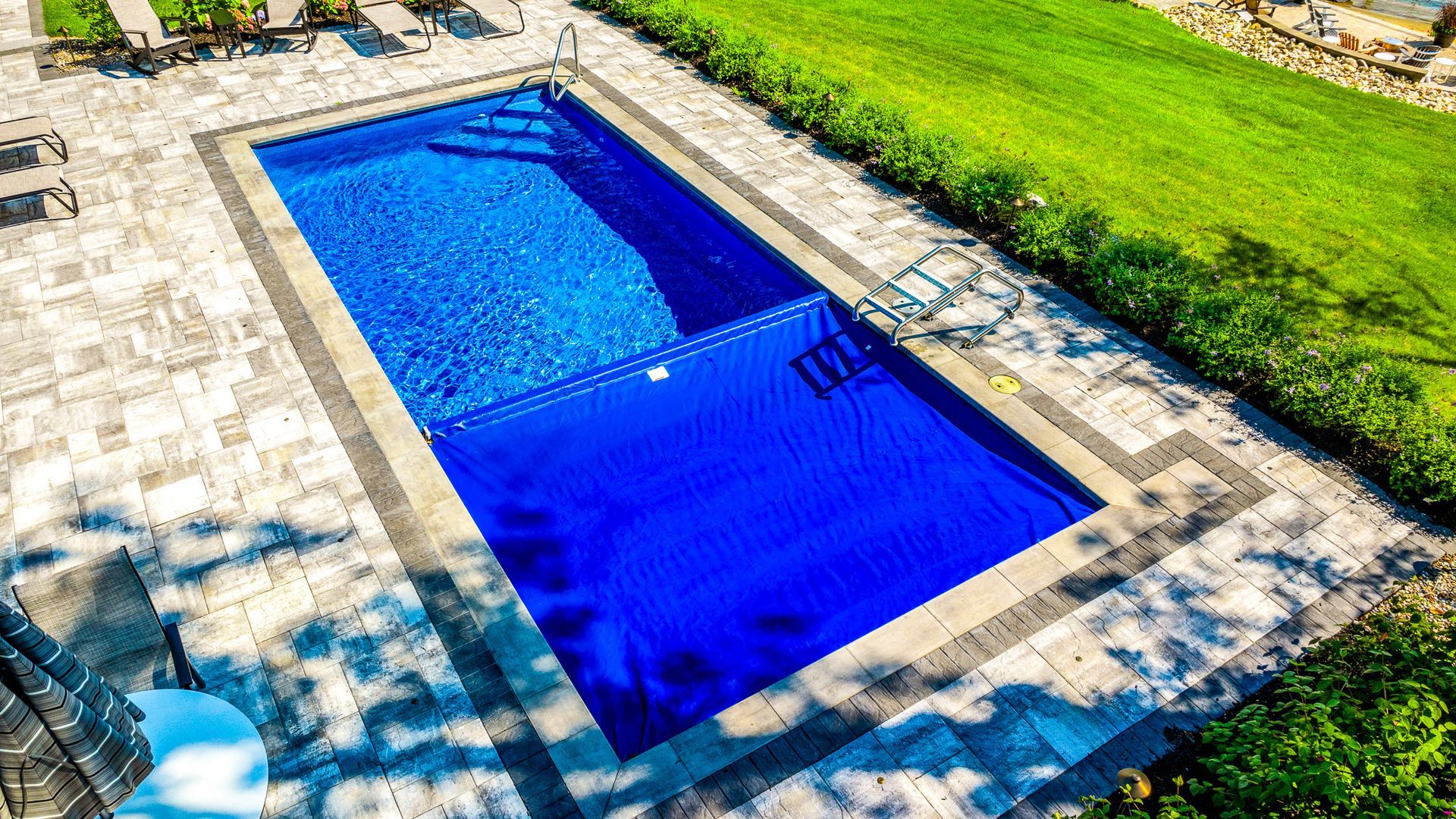 Rectangular blue swimming pool surrounded by stone paving and green grass in bright sunlight