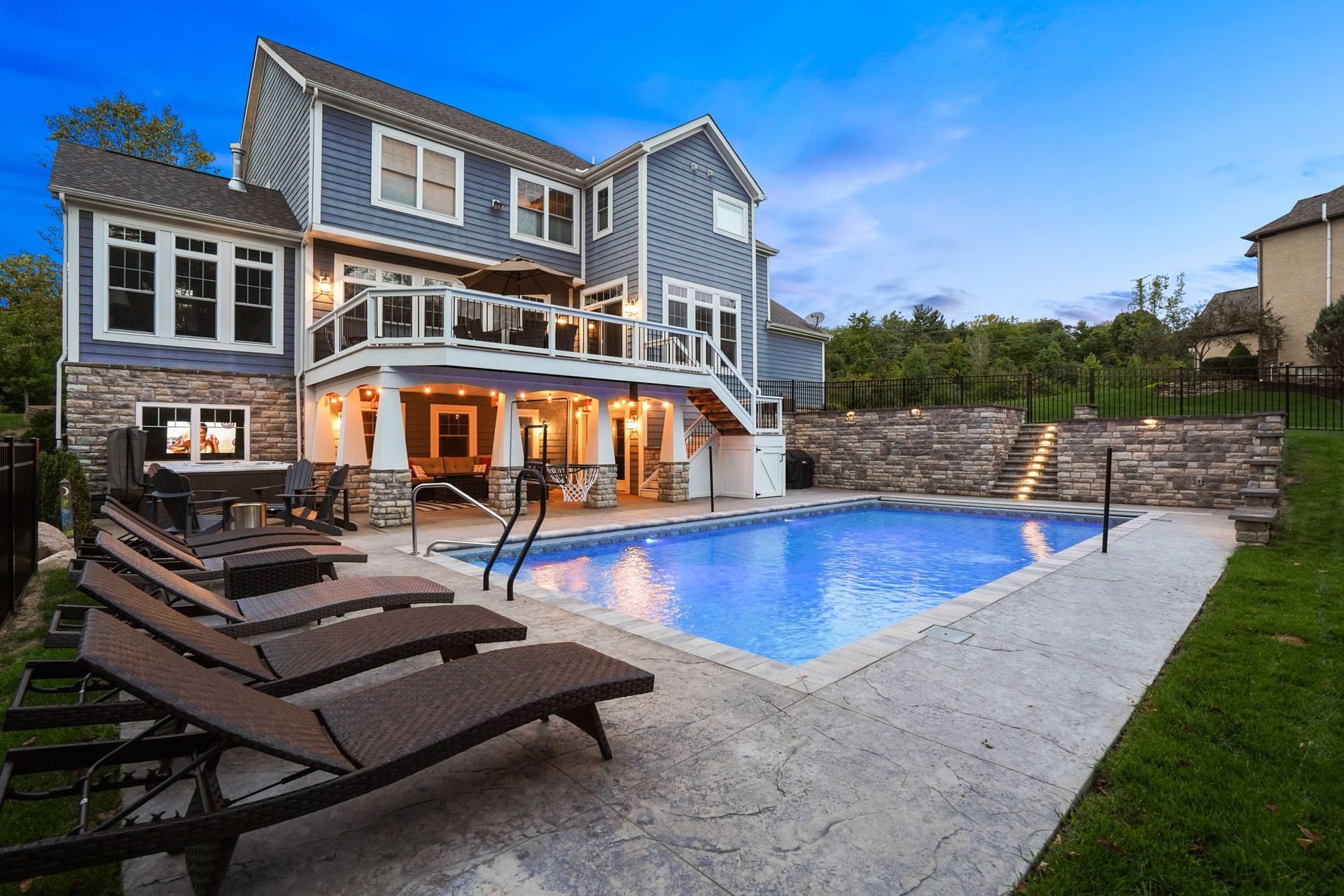 Modern two-story house with a lit backyard pool, patio seating, and stone retaining wall at dusk