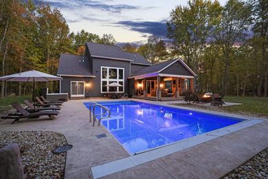 Modern house with lit blue pool, lounge chairs, and wooded backyard at dusk