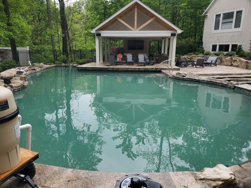 Backyard pool with turquoise water, stone edging, and a covered patio beside a house and trees
