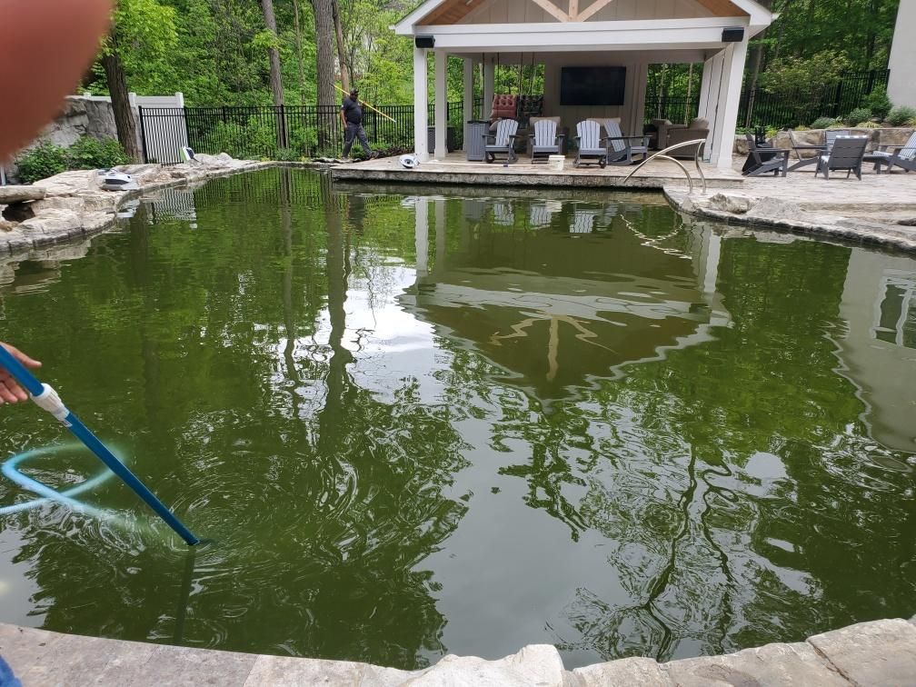 Green pond with a pavilion and lounge chairs by the water, reflecting trees and the sky.