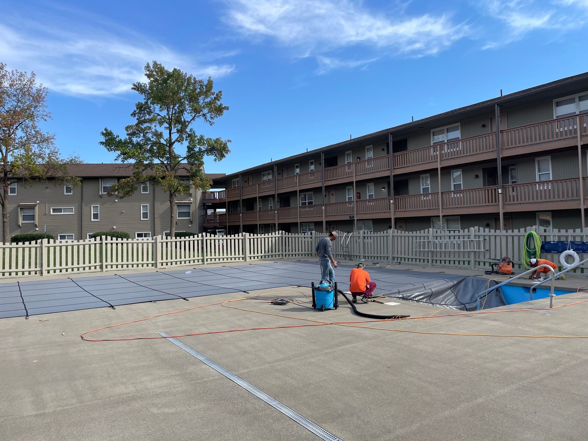 Outdoor apartment courtyard with workers, a fence, tarps, and a long brick building under a blue sky