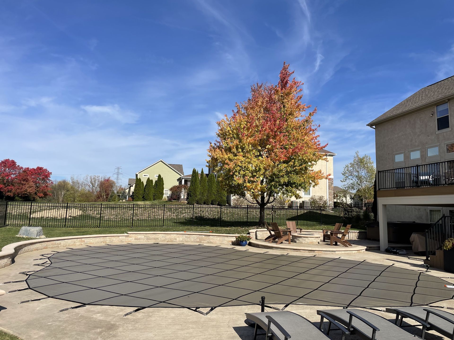 Sunlit courtyard with a large tree, stone patio, benches, and residential buildings under a blue sky.