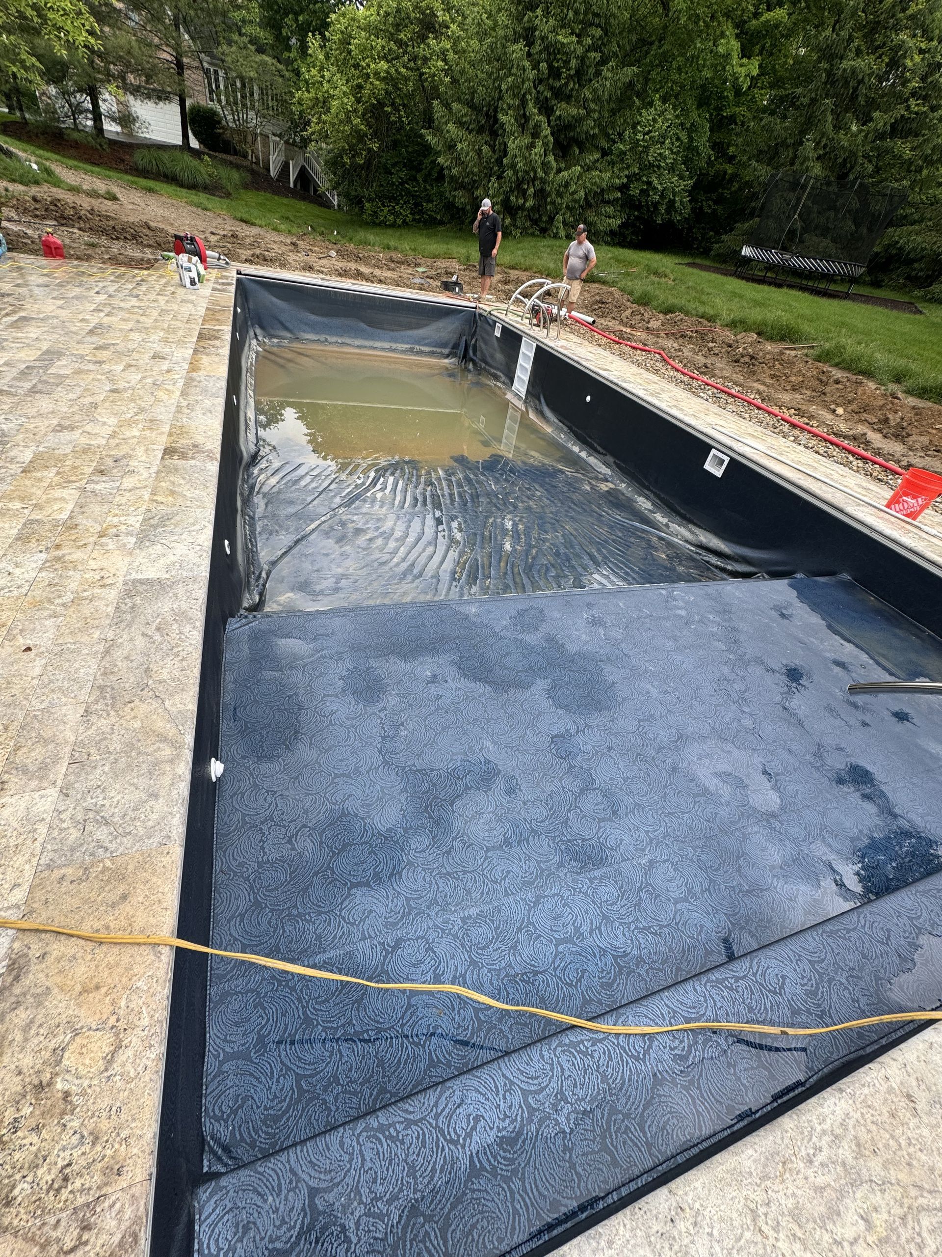Rectangular outdoor pool under construction with black liner, gravel edge, and caution tape nearby