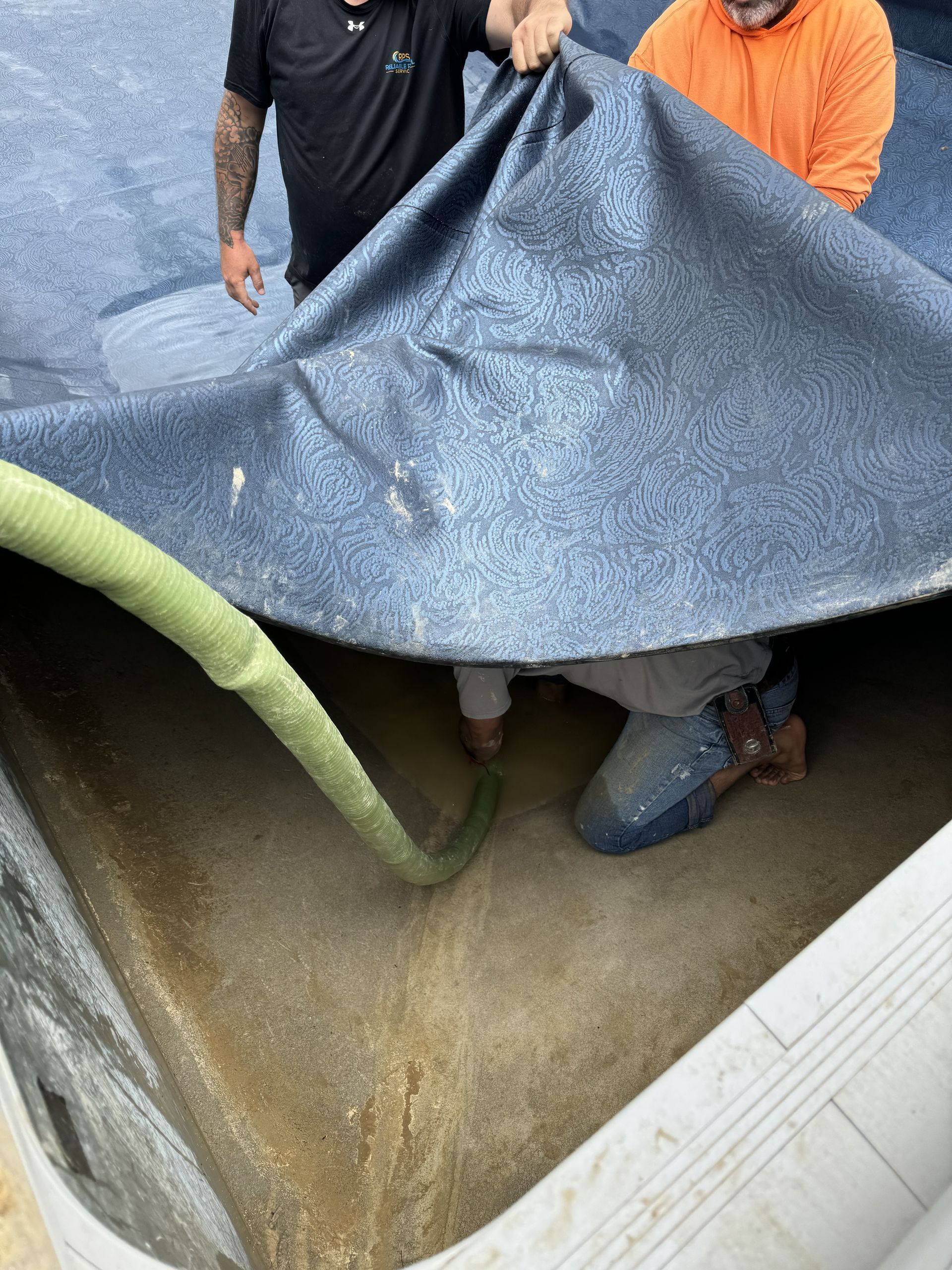Workers lifting a tarp over a flooded pit with muddy water and pipes exposed