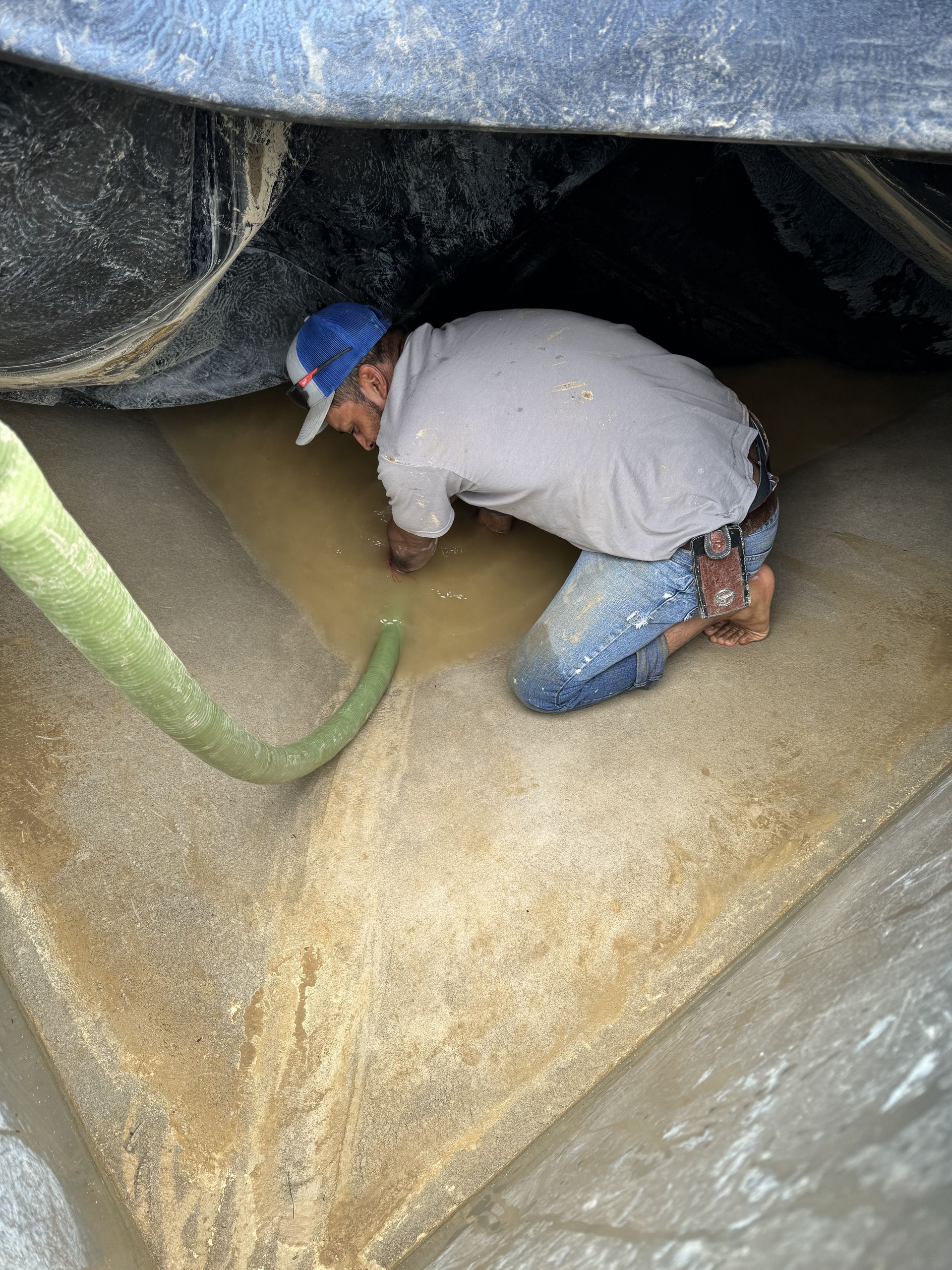 Worker in blue hard hat crouches inside a grain silo, inspecting piled grain near a green auger.