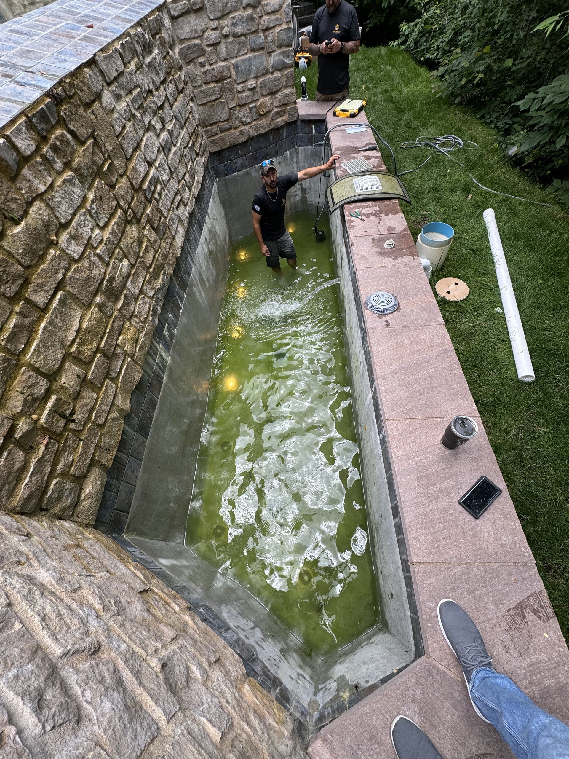 Two people in a narrow stone water channel, one wading in green water beside a paved walkway.