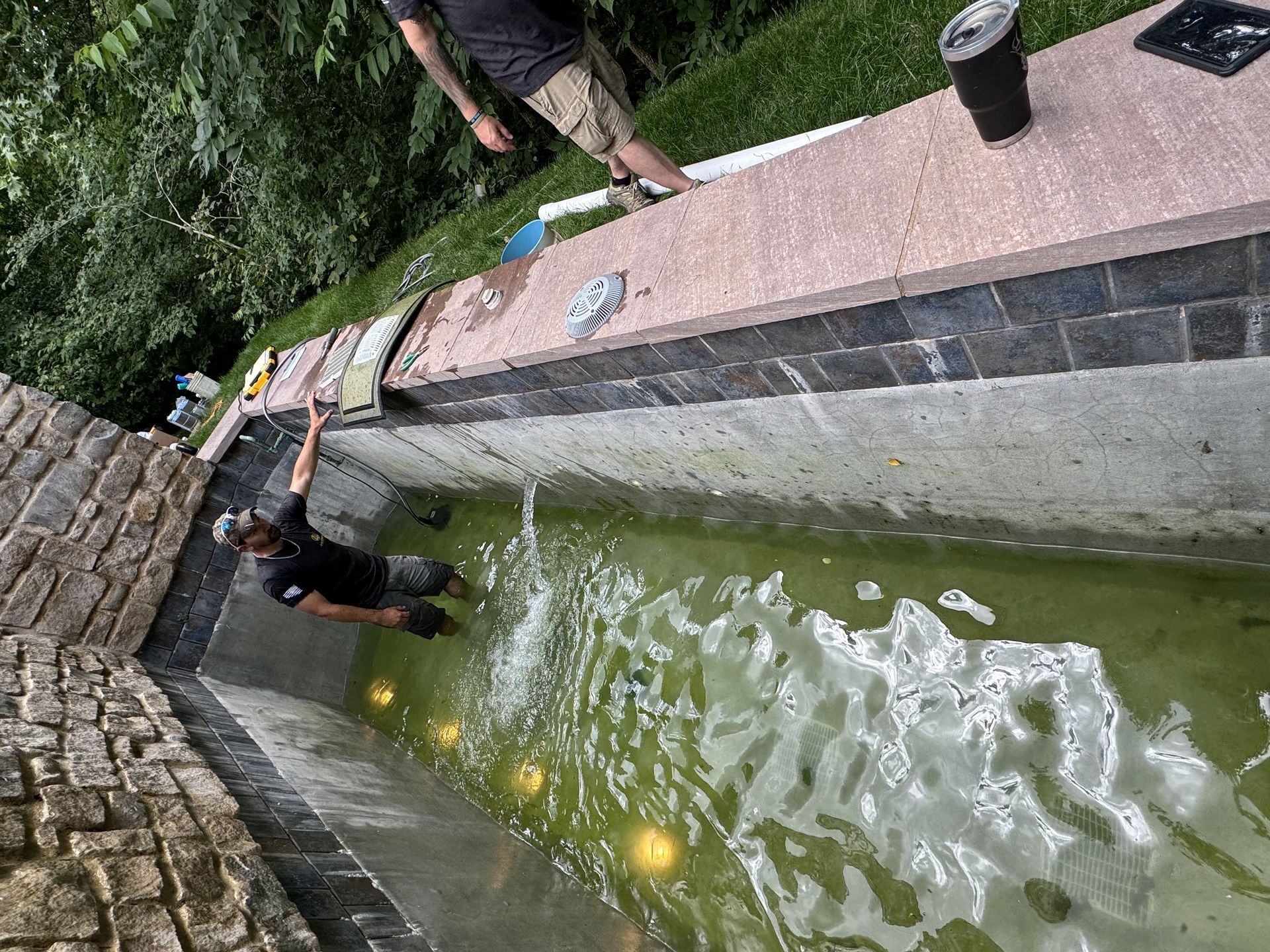 Person leaning over a narrow green canal beside a stone wall and walkway with a cup on the ledge