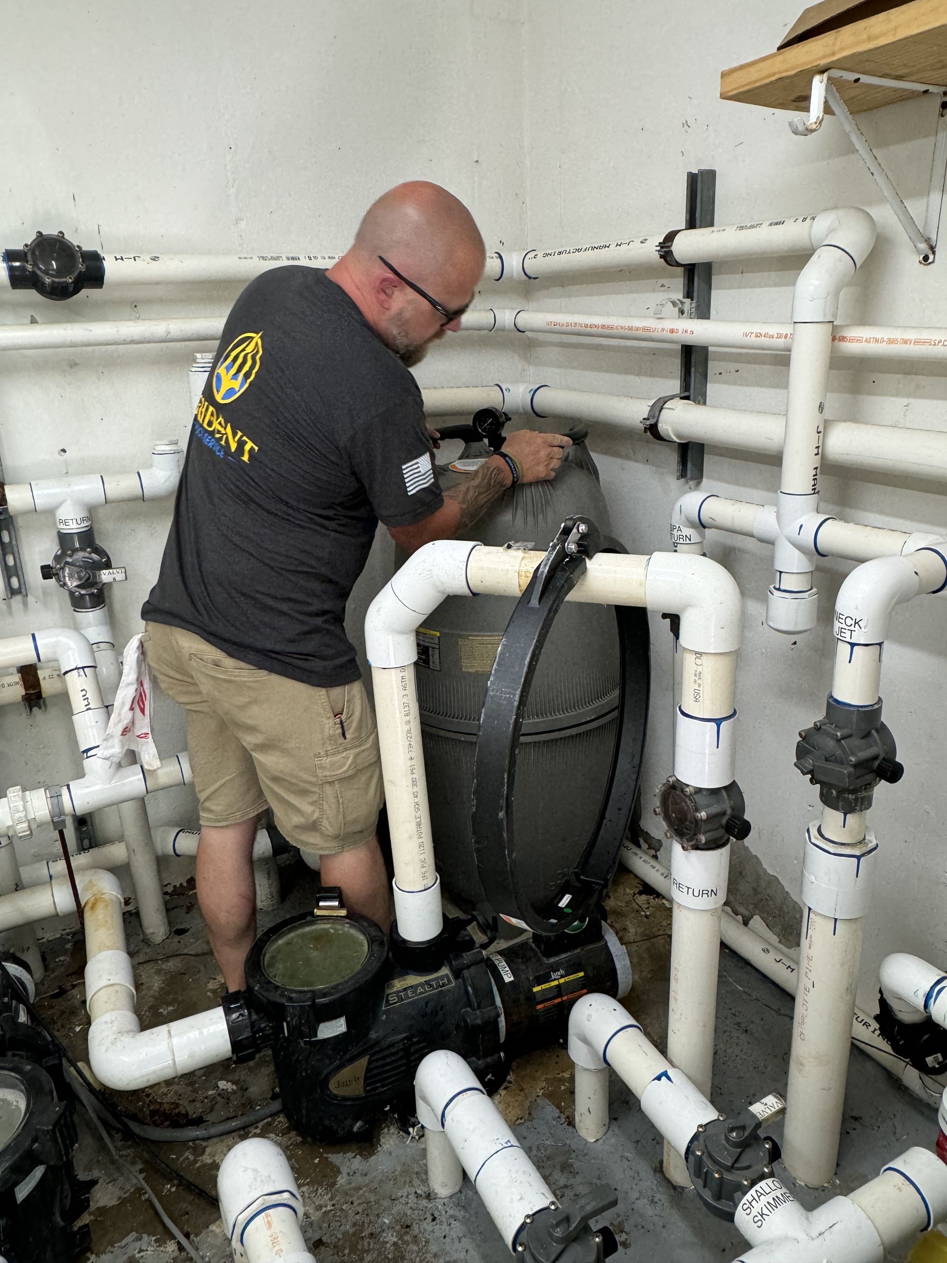 Man in black shirt and khaki shorts working on white PVC pipes and equipment in a utility room