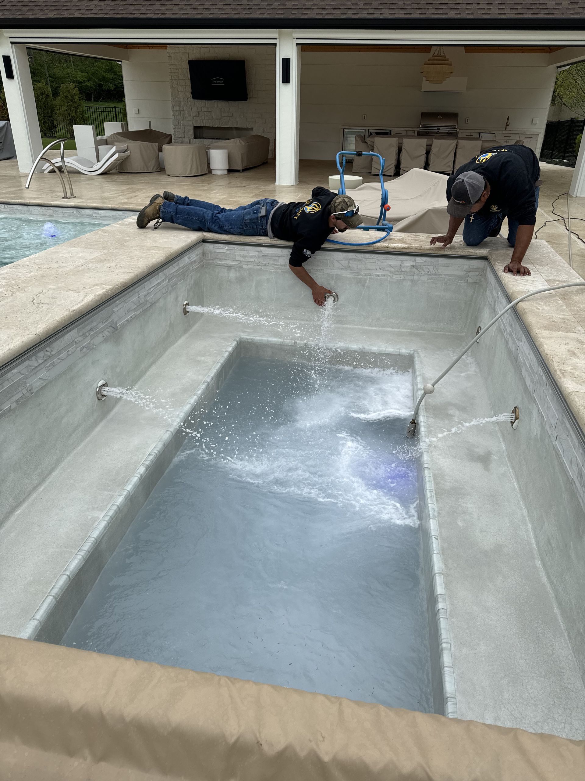 Two workers inspecting an empty concrete pool beside a patio and covered outdoor seating area