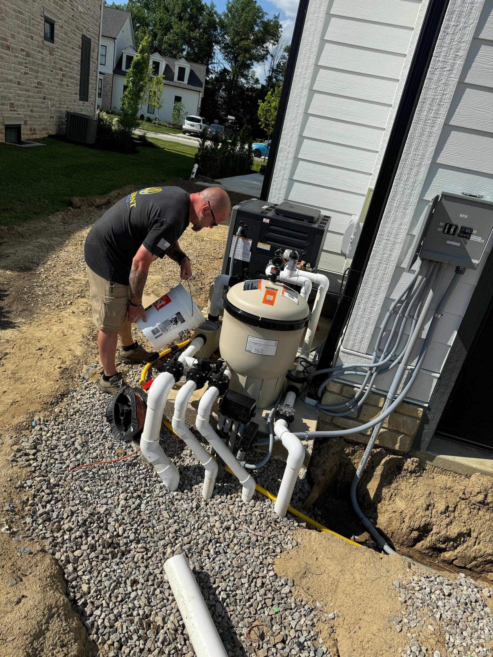 Worker servicing pool equipment beside a house, with pipes and hoses on gravel.