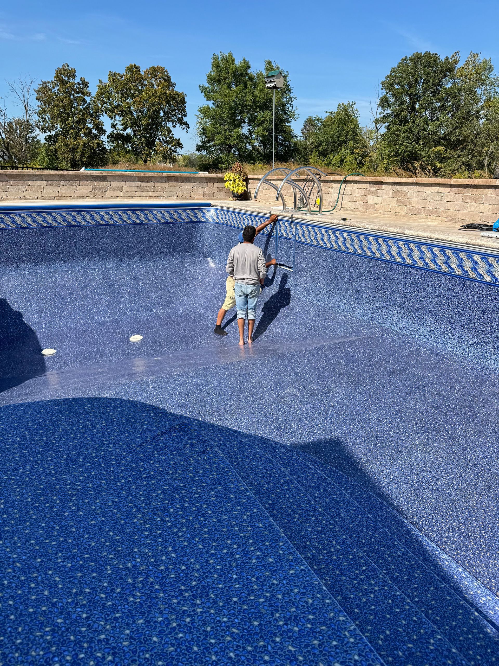 Worker inside a blue in-ground pool, cleaning or repairing it under a sunny sky.