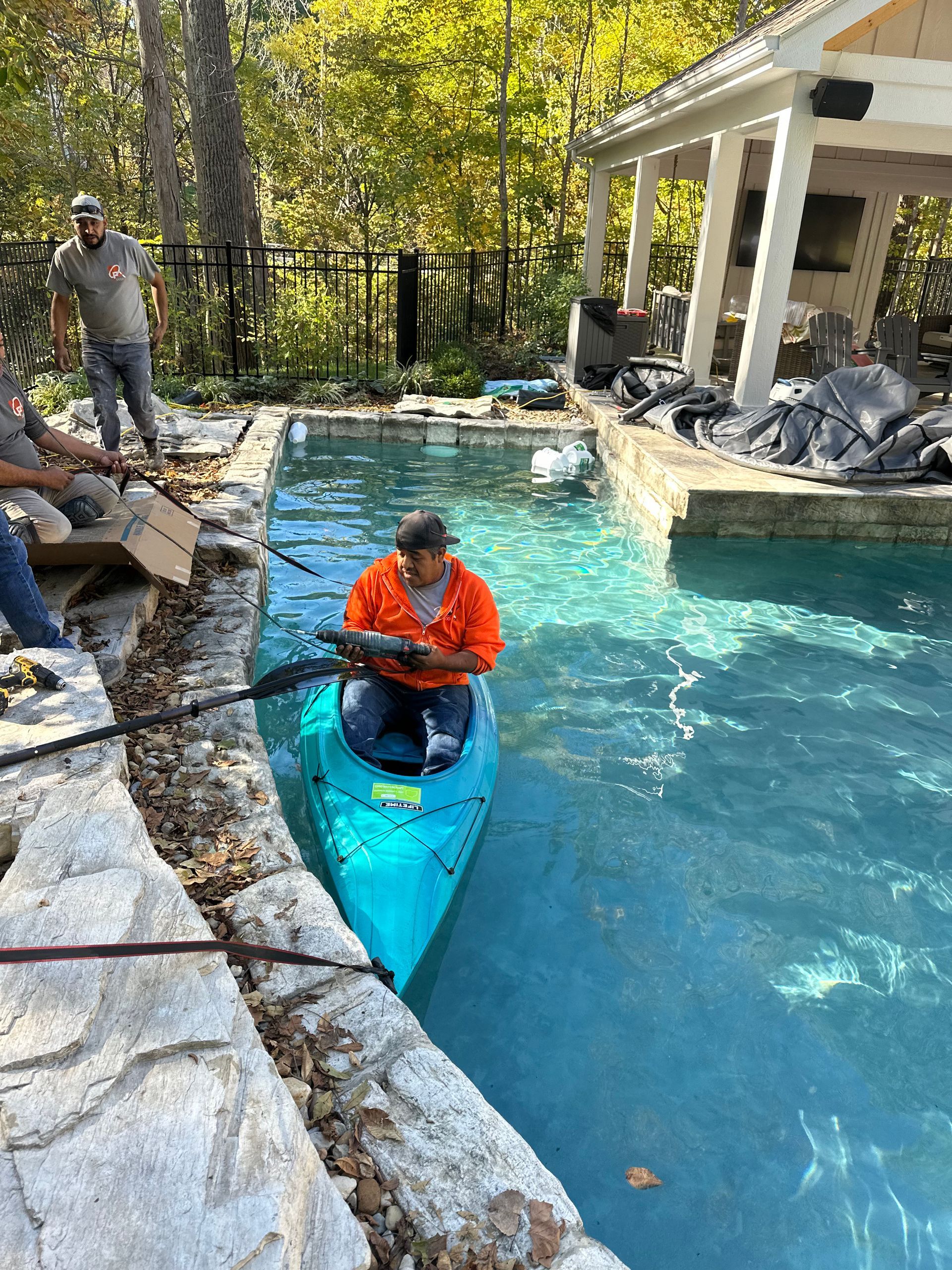 Person in an orange jacket sits in a blue kayak in a backyard pool with people nearby.