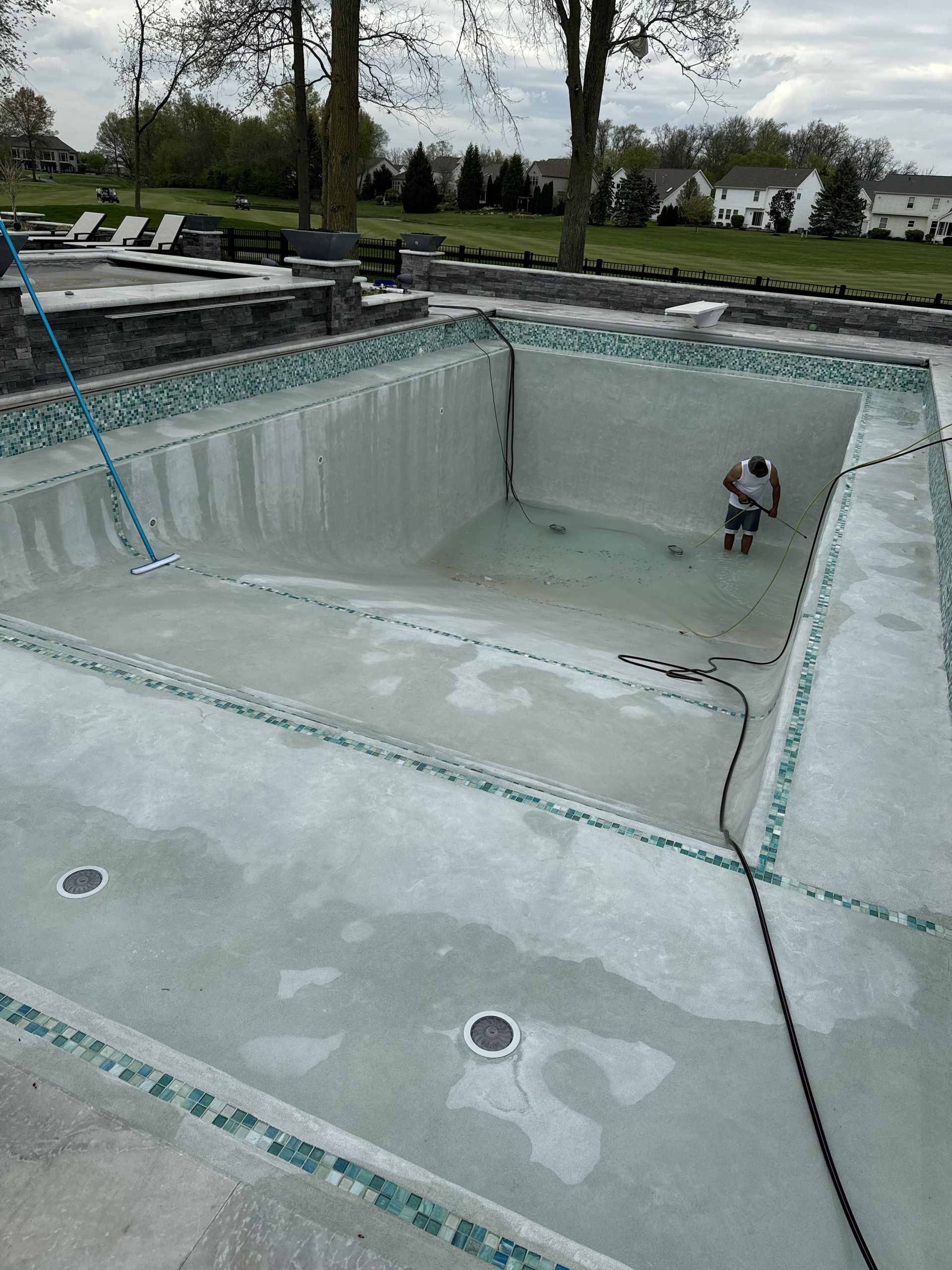 Empty drained swimming pool being cleaned, with a person inside and a hose on the edge.