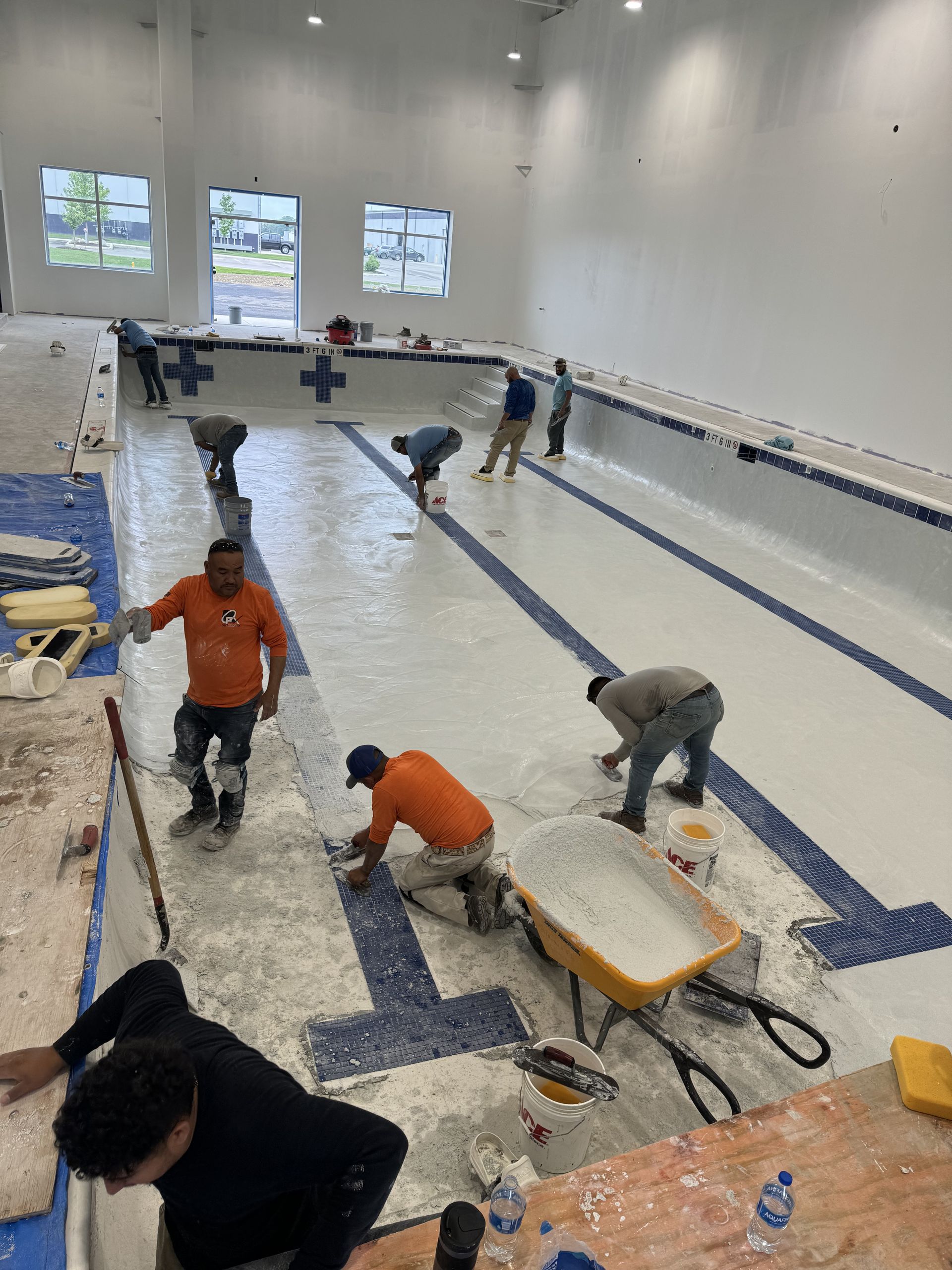 Workers repairing a drained indoor pool, with tools, buckets, and blue tile lines visible.