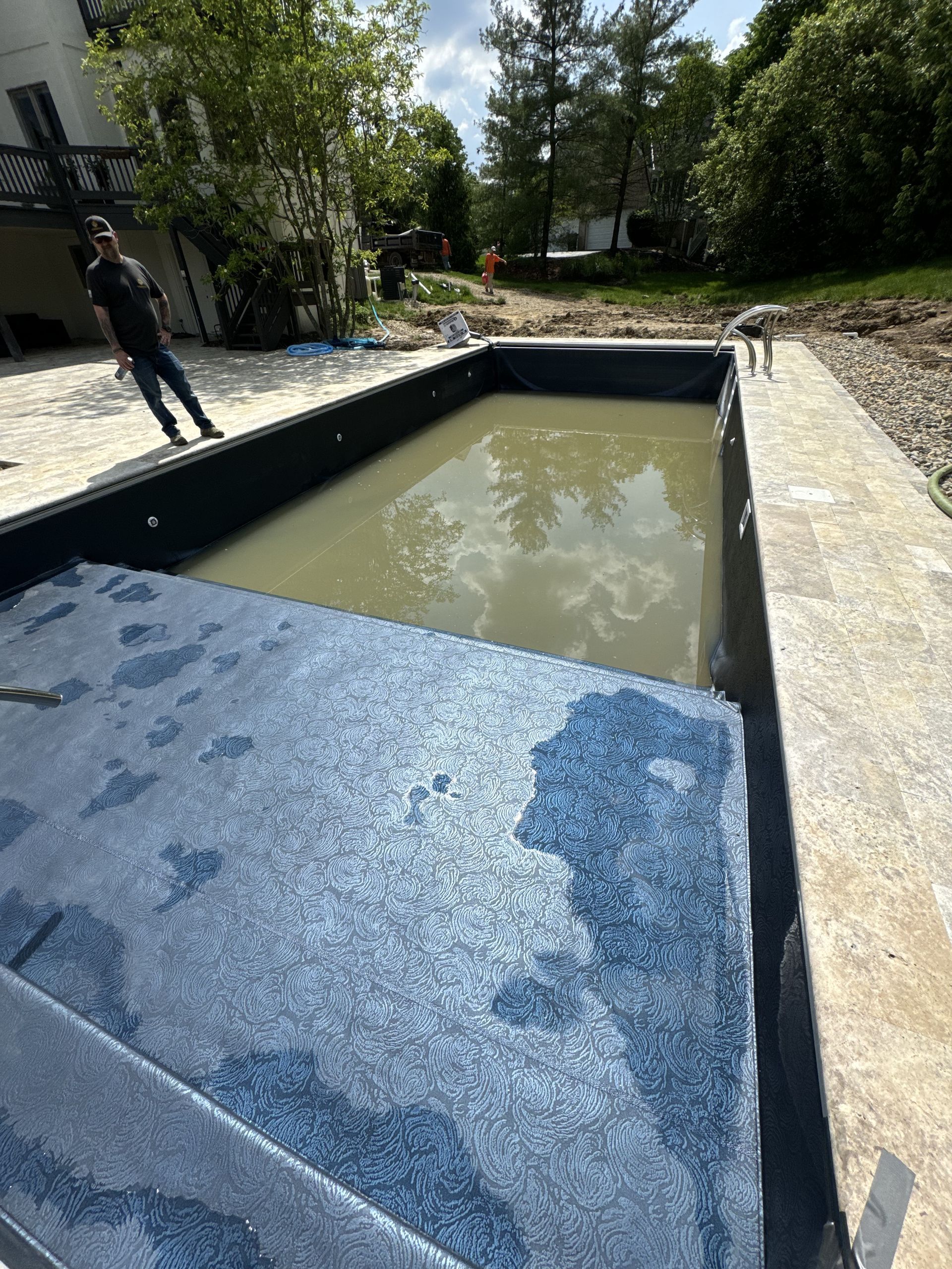 Rectangular backyard pool under construction, with a blue liner and muddy water, surrounded by gravel and trees