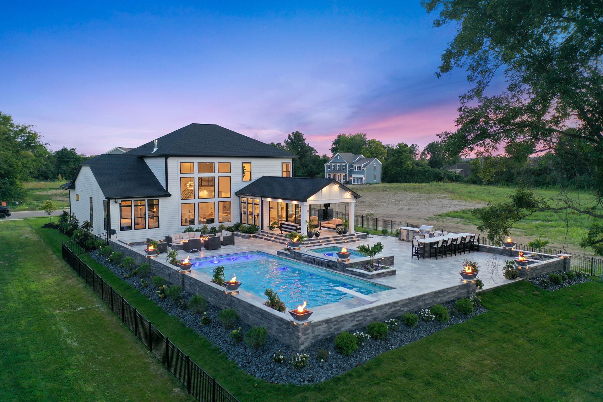 Modern house with a lit backyard pool and patio at dusk, surrounded by a stone wall and green lawn