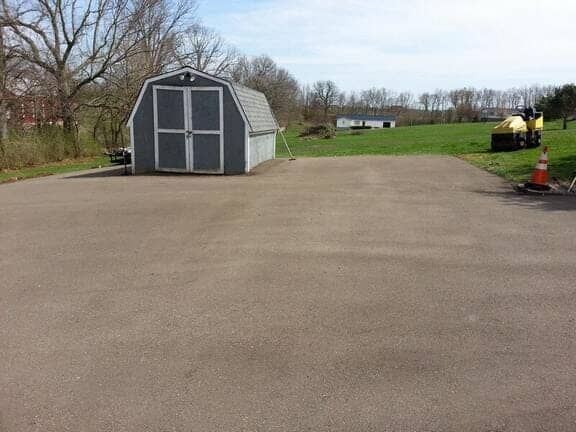 A grey shed sits in the corner of a large, paved parking lot on a sunny day, with a yellow construction vehicle nearby.