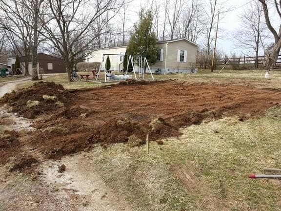 A yard showing an area of cleared, reddish-brown soil prepared for construction in front of a mobile home.