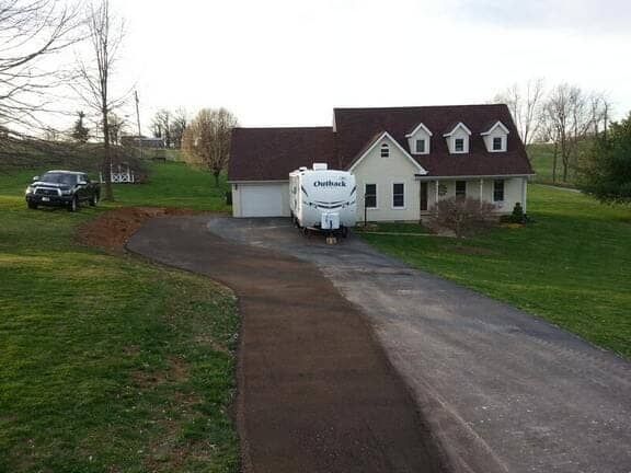 A beige house with a dark roof and a camper parked in the driveway, with a black truck parked on the lawn to the left.
