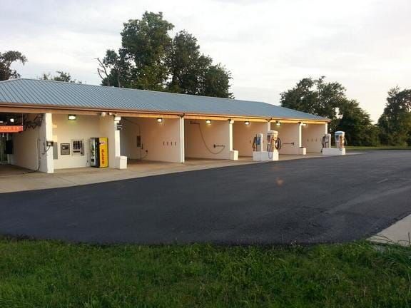 A self-service car wash facility with a light blue metal roof, white walls, and empty bays under a bright, cloudy sky.