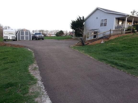 A paved driveway leads to a light blue house, a shed, a parked truck, and a trailer on a grassy lot.