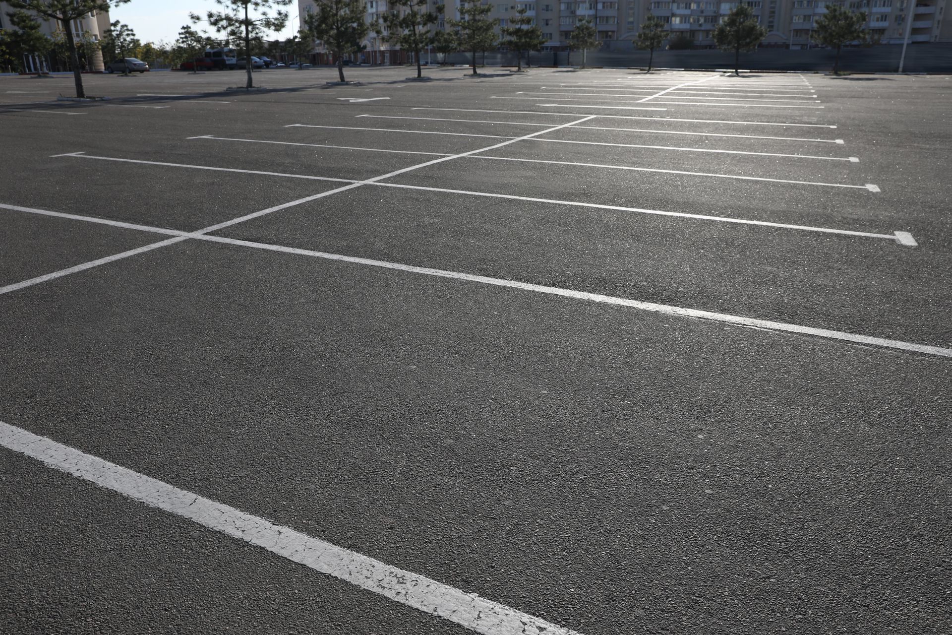An empty outdoor asphalt parking lot with painted white parking space lines under a bright sky. An empty outdoor asphalt parking lot with painted white parking space lines under a bright sky.