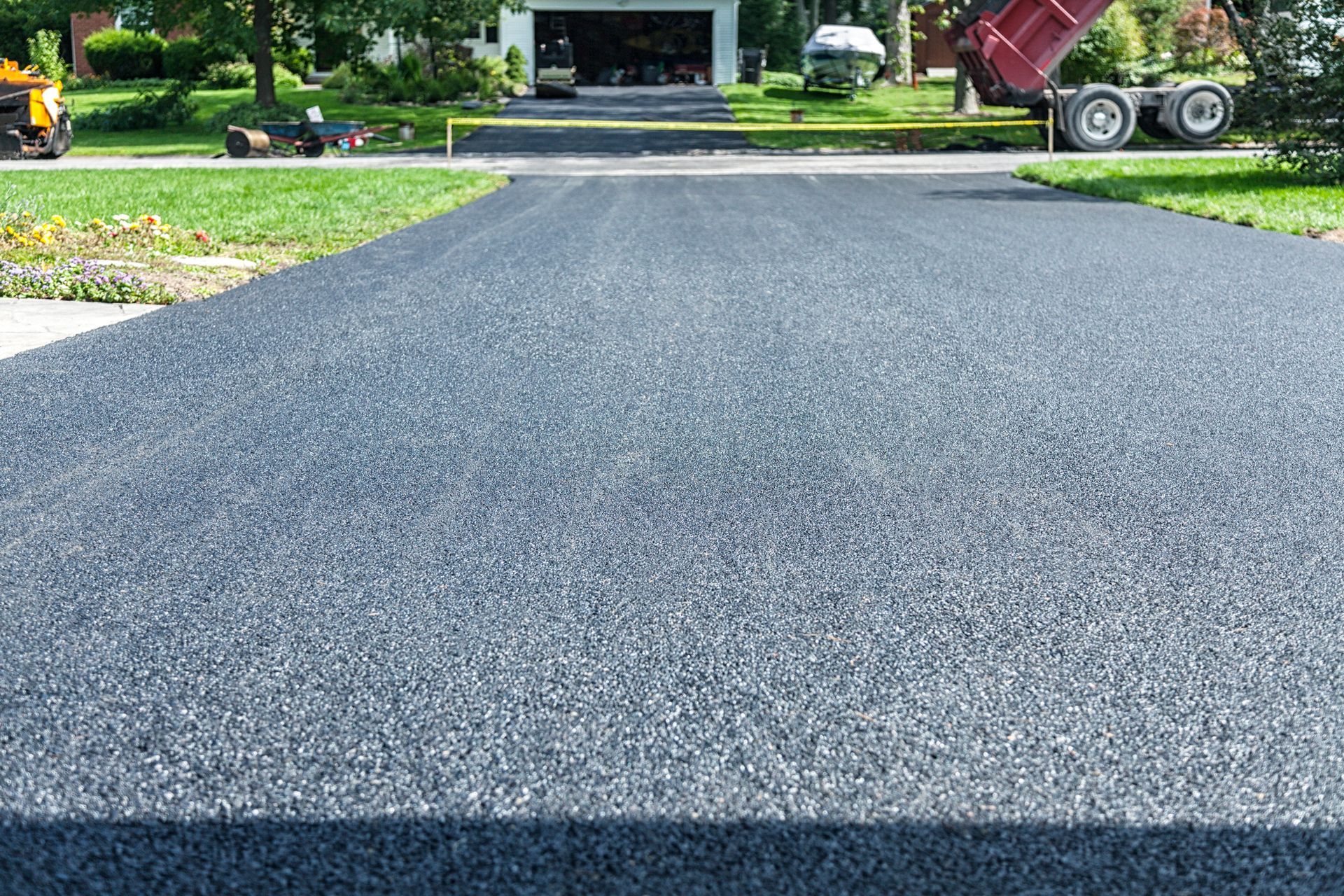 A newly paved, dark asphalt driveway leading toward a residential garage on a sunny day, with construction equipment nearby.