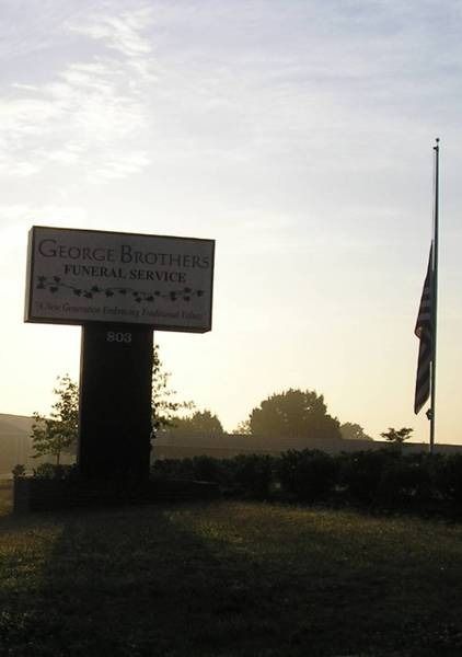 Sign for George Brothers Funeral Service with flag at half-mast under a bright sky.