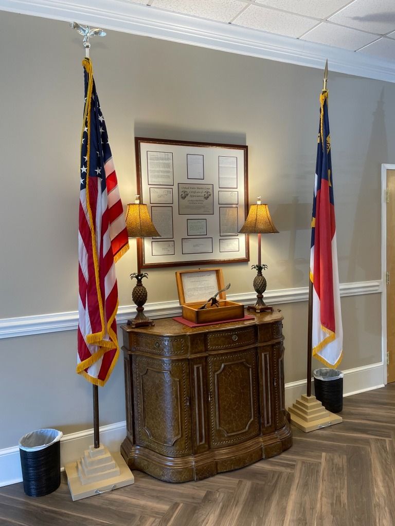 Flags flank a wooden cabinet with lamps and documents, North Carolina and US flags displayed indoors.