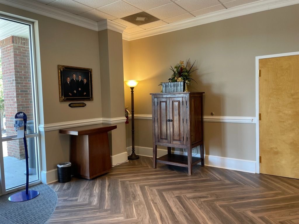 Lobby with wooden floors, cabinet, portrait, and a door. There is a lamp and an entrance. Beige walls.