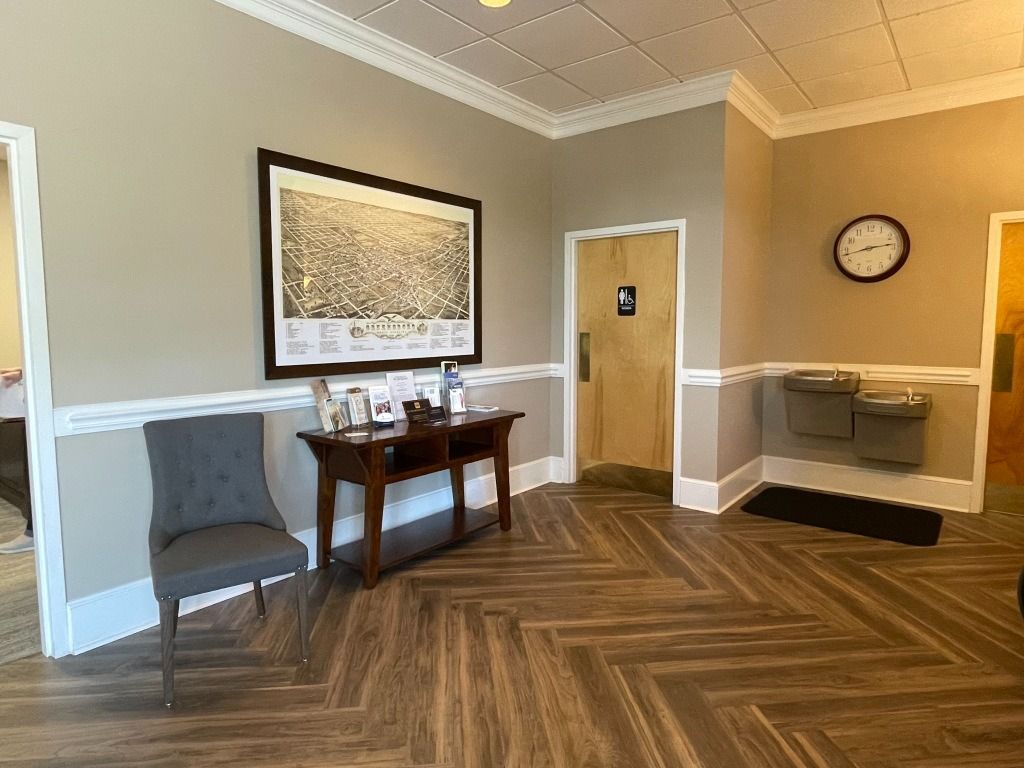 Reception area with a desk, chair, clock, and water fountain. Herringbone wood floor, beige walls, and white trim.