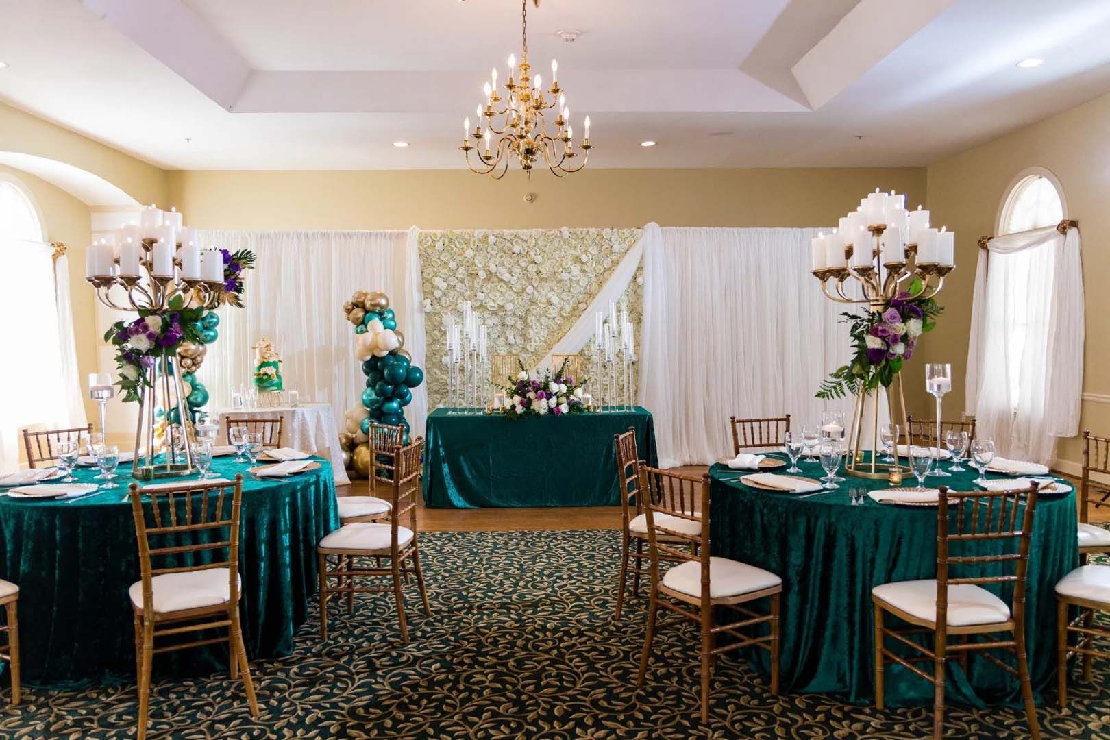 Elegant event room with teal tablecloths, gold chairs, large floral centerpieces, and a floral backdrop.