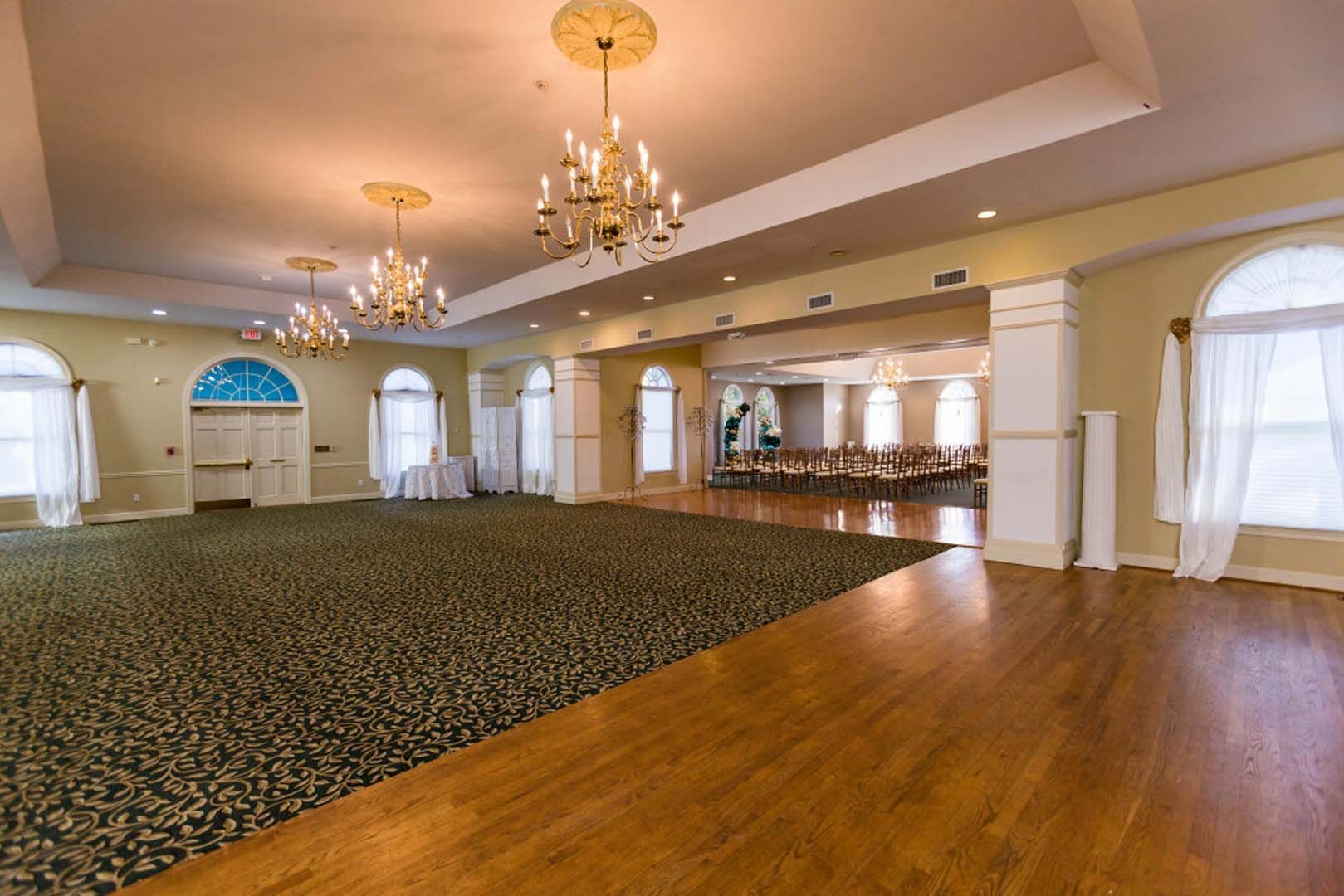 Ballroom with green carpet and wood floor, chandeliers, arched windows, and a doorway to another room.