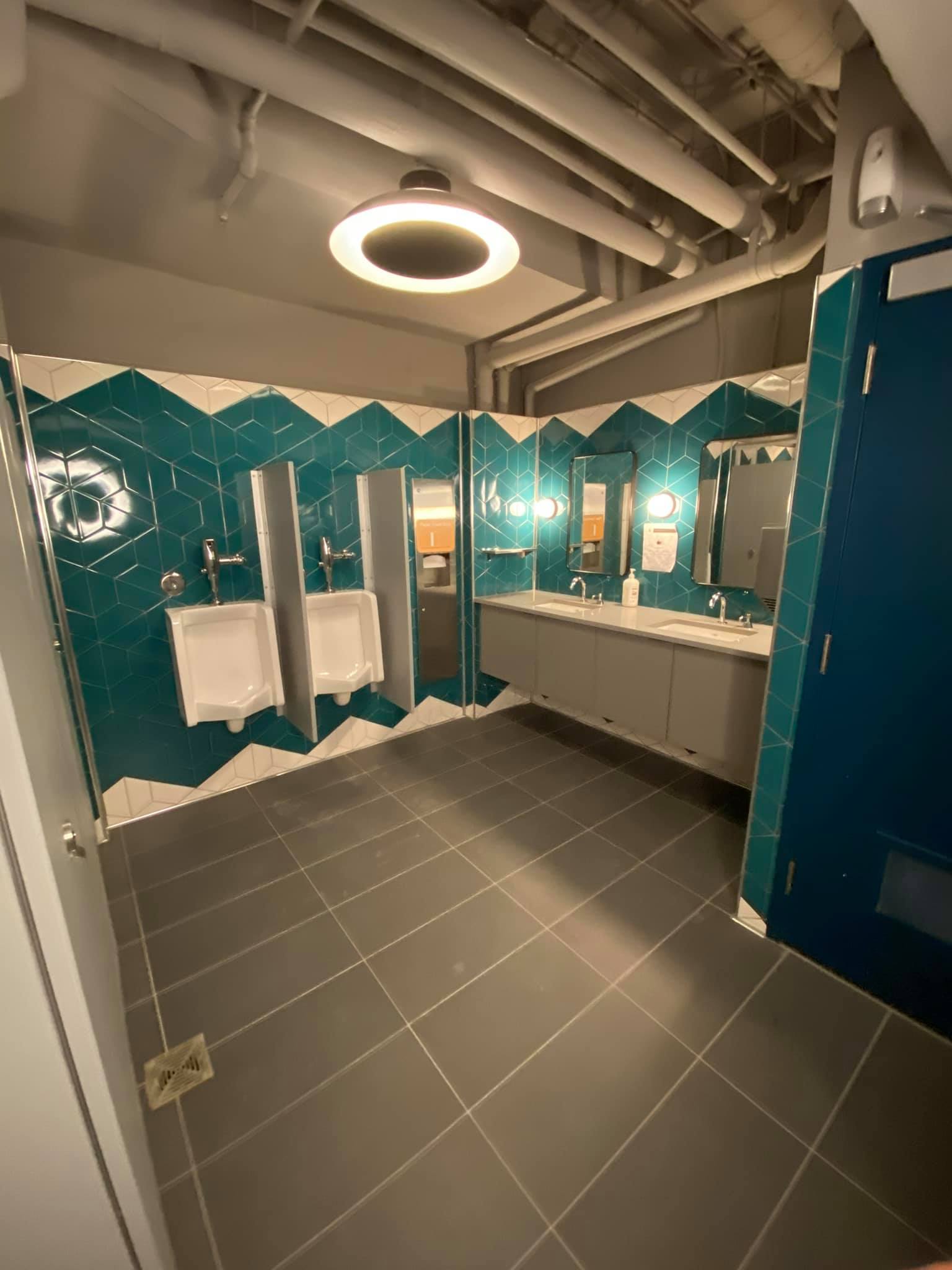 Modern restroom with teal tiled walls, white sinks, urinals, mirrors, and dark gray floor tiles