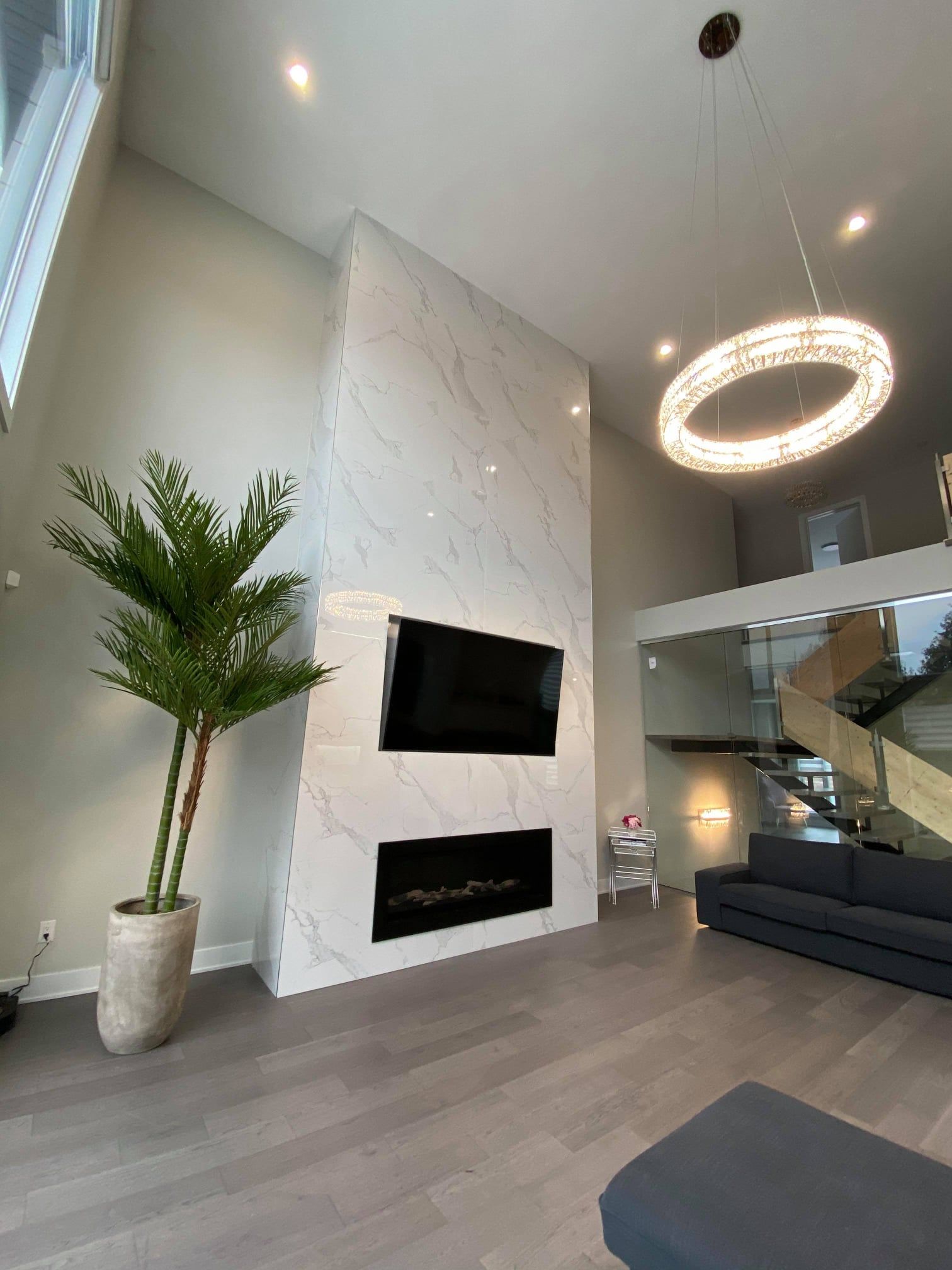 Modern living room with marble fireplace, floating stairs, large ring chandelier, and dark sectional sofa.