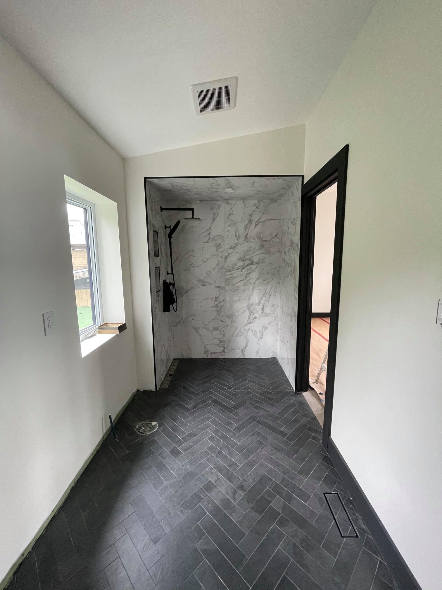 Narrow hallway with dark tile floor, white walls, marble shower area, window, and open doorway.