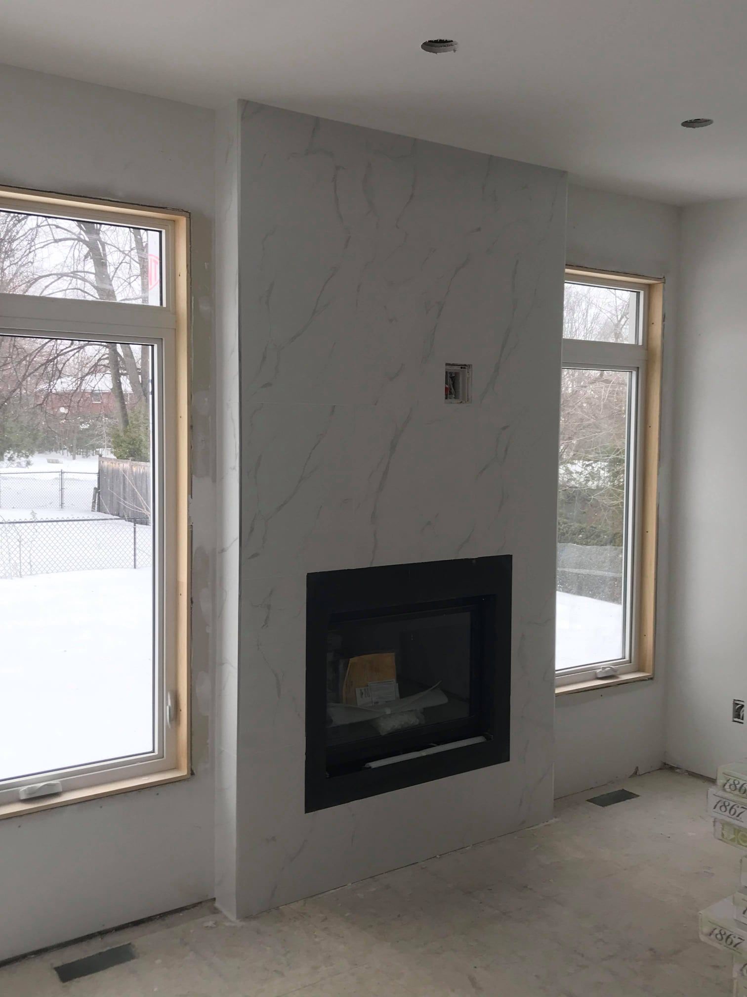 Modern living room with a gray fireplace wall and two tall windows, unfinished floor.