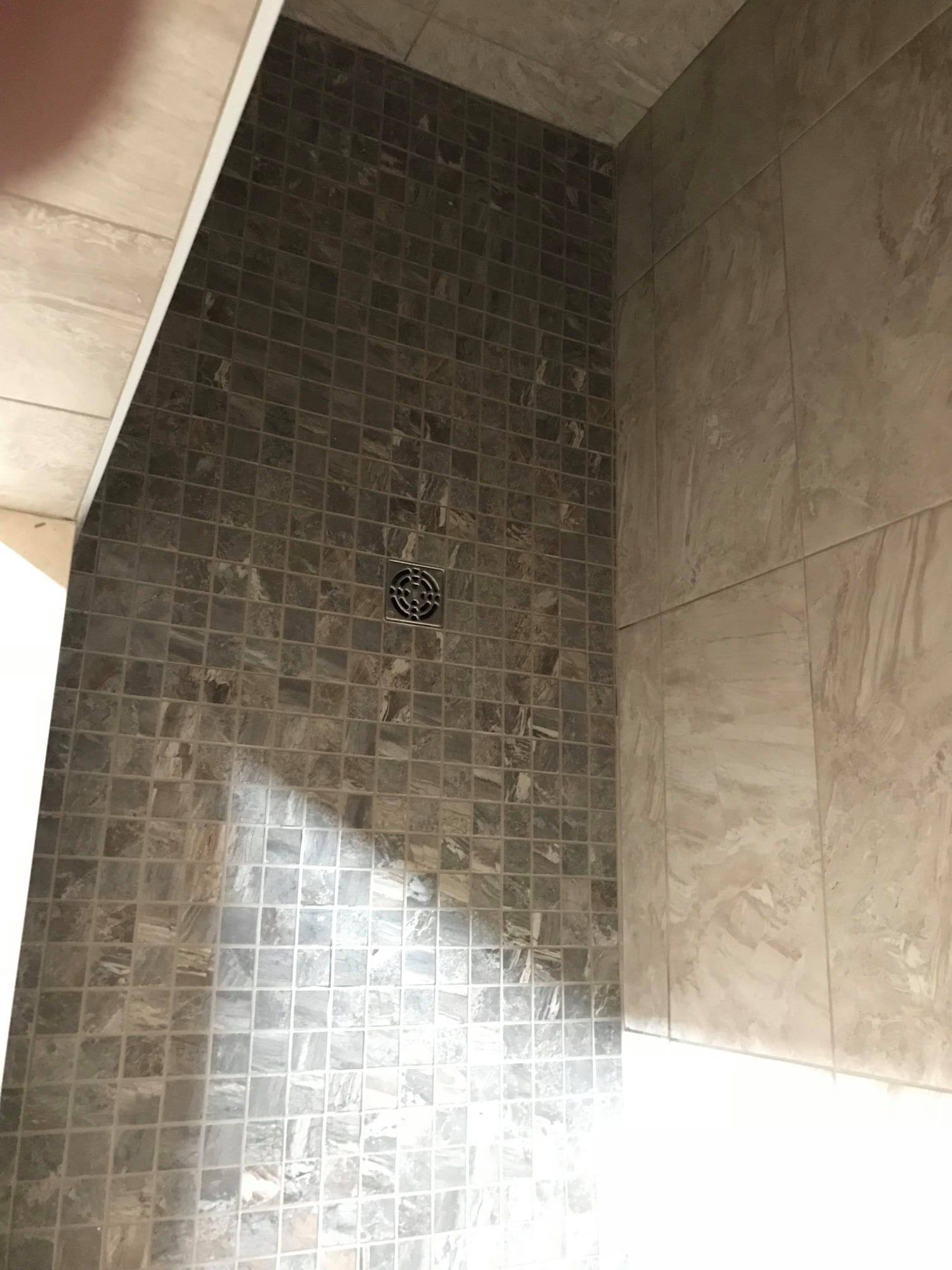 Tiled shower wall with a small square drain, beige stone walls, and sunlight falling across the floor.