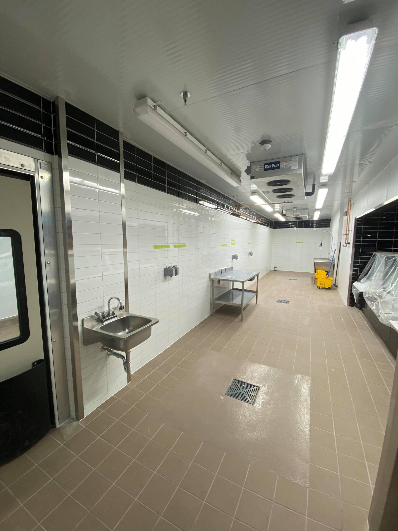 Long tiled corridor with white walls, sinks, benches, and overhead fluorescent lights, resembling a locker room or station hallway