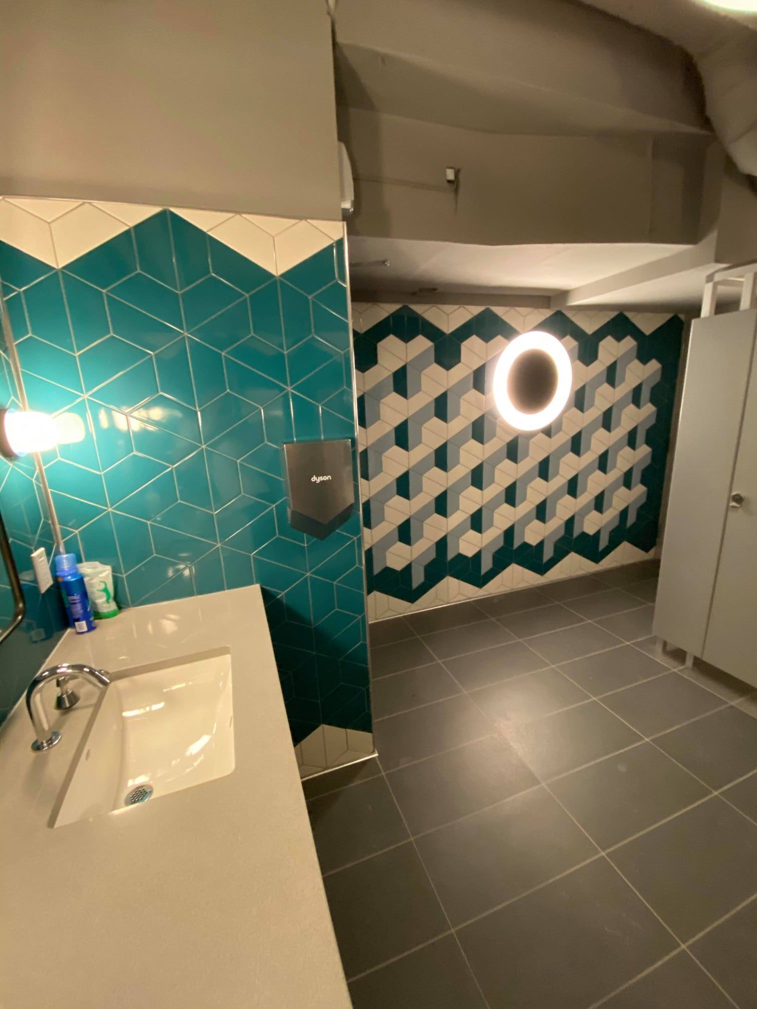 Modern bathroom with teal tile walls, white sink, gray floor, and lit round mirror above a patterned partition