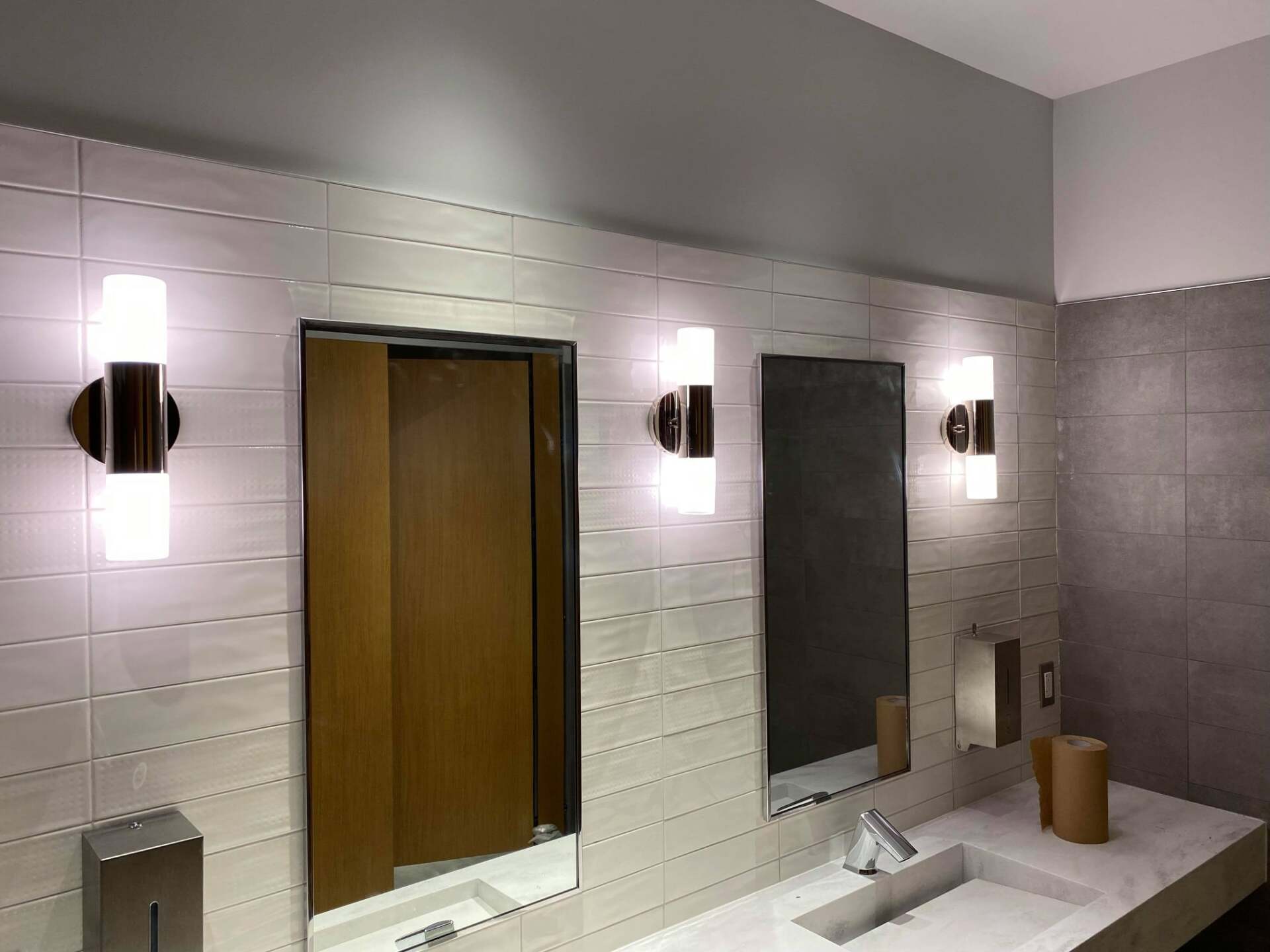 Public restroom with three mirrors, wall sconces, sinks, and a wooden toilet stall door