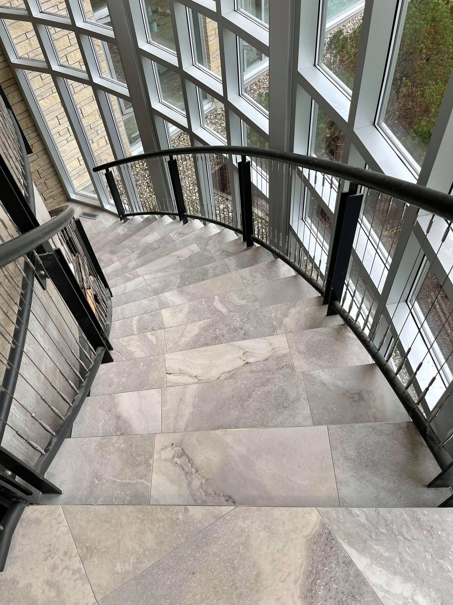Spiral staircase with stone steps and black railing beside large windows.